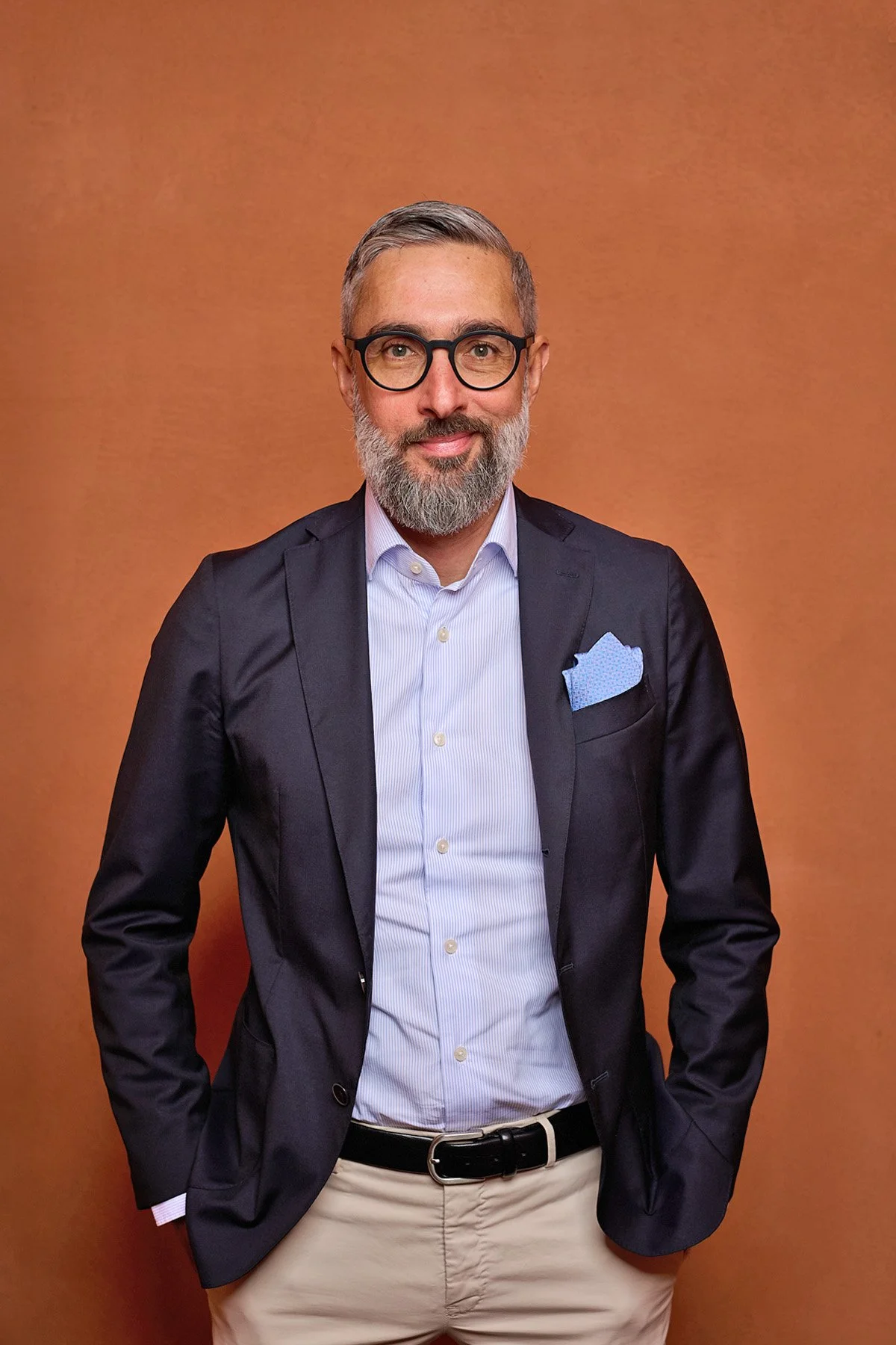 A man with gray hair, glasses, and a beard wearing a dark blazer, light blue shirt, and beige pants standing against a plain orange background.