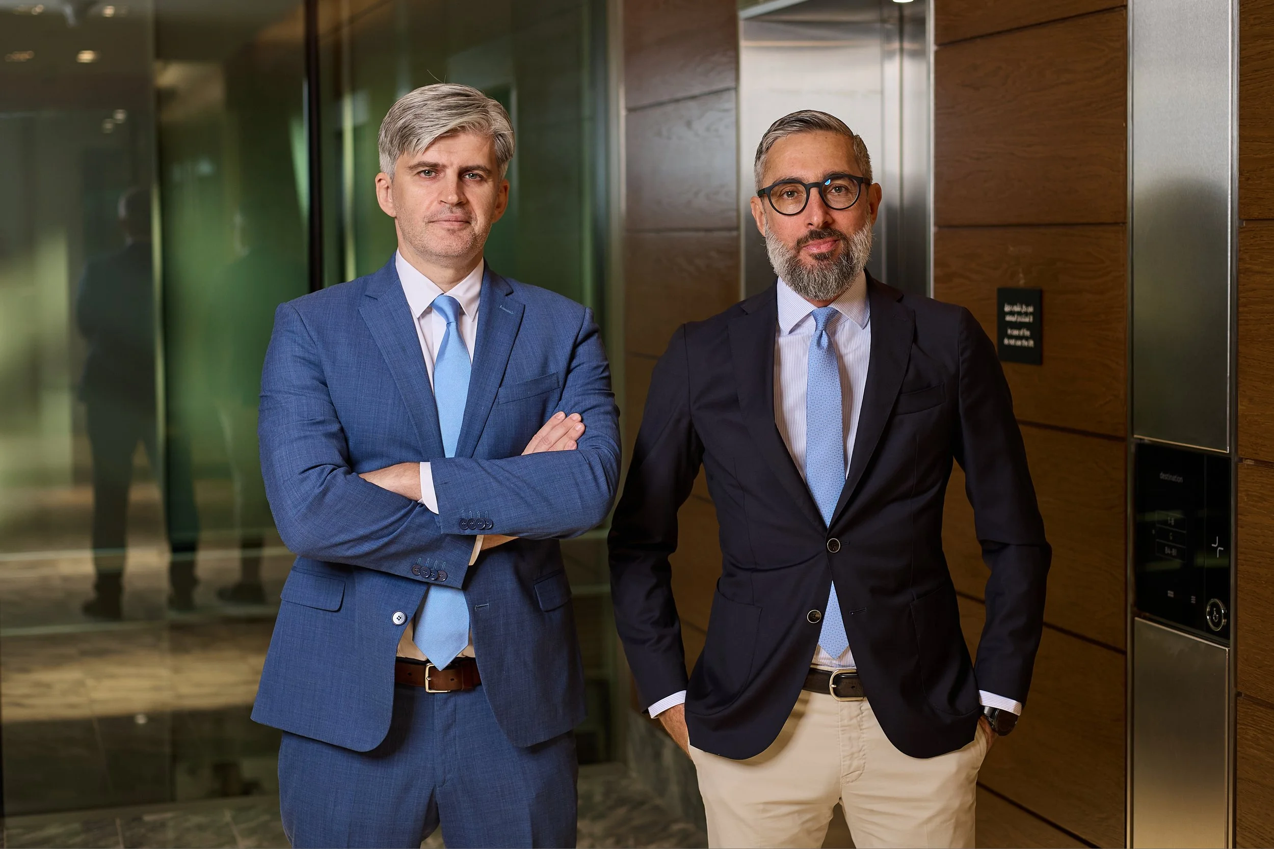 Two professionally dressed men standing in front of an elevator in an office building lobby.