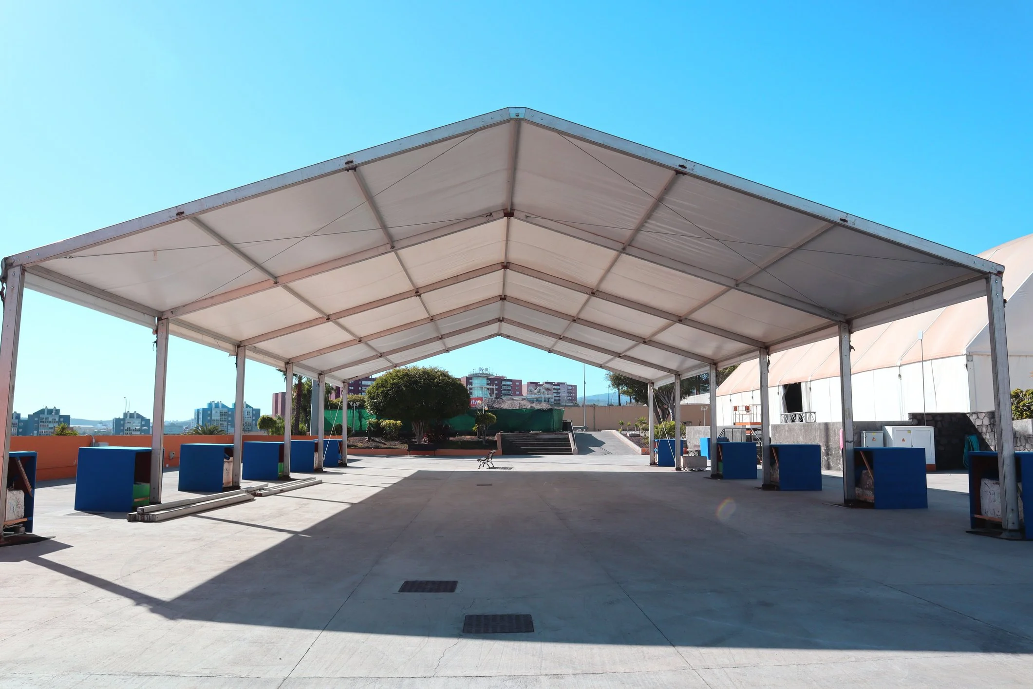 Empty outdoor event tent on concrete with blue amplifier boxes, trees, and buildings in the background under a clear blue sky.