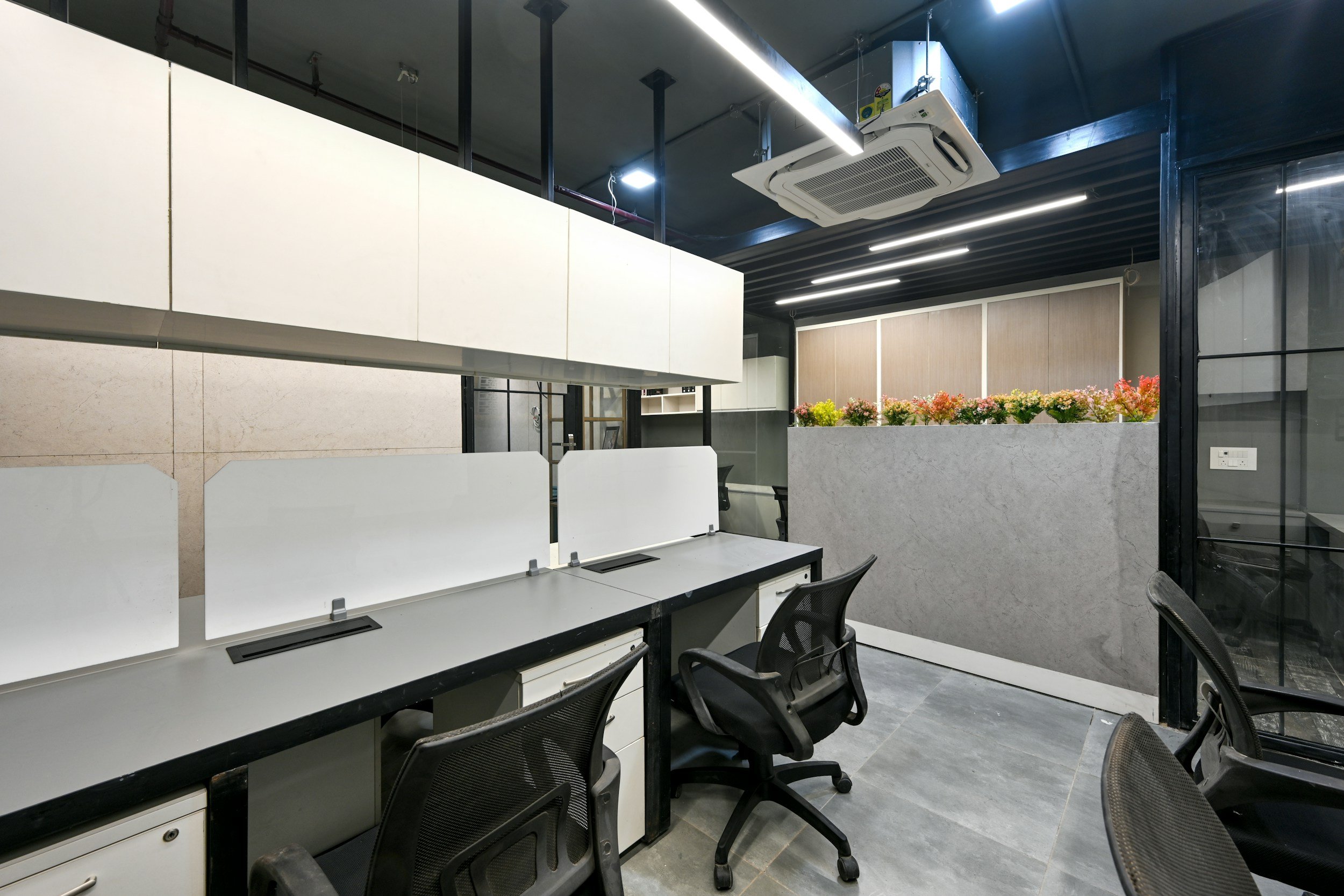 Modern office workspace with partitioned desks, black office chairs, a flower planter, ceiling air conditioner, and black ceiling beams.