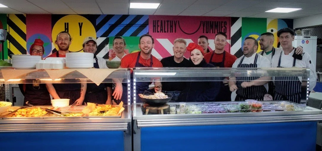 Group of chefs and staff smiling behind a food display counter in a colorful kitchen with a mural background.