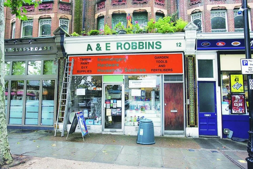 The storefront of A & E Robbins, a hardware and household supplies store, with a large orange sign listing items like timber, paint, DIY supplies, tools, and fertilizers. The store has glass windows with various signs and advertisements. A ladder lea