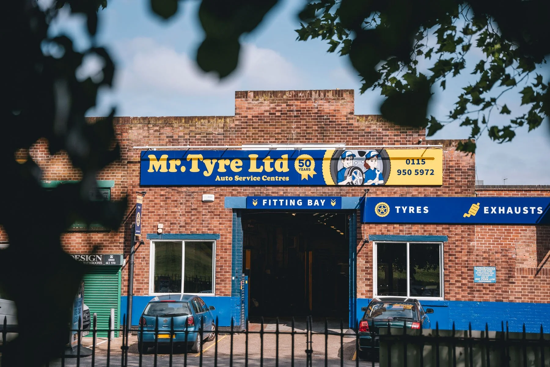 Braided brick building with a sign reading 'Mr. Tyre Ltd. Auto Service Centres'. The sign features cartoon characters of a man and a woman wearing mechanic uniforms. Labels on the building indicate it is a fitting bay for tyres and exhausts, with par