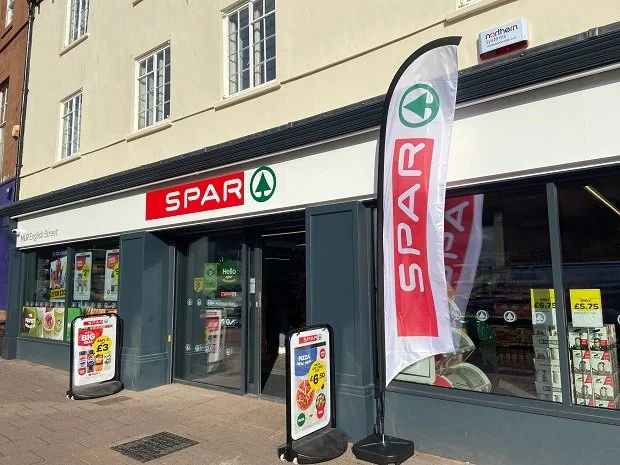 Exterior of a SPAR grocery store with promotional signs outside and a red and white SPAR flag.