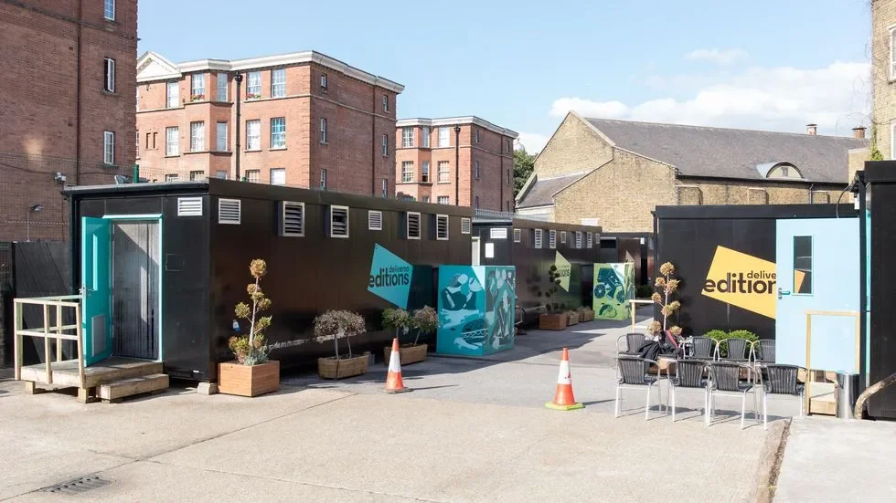 A rooftop area with black containers that have colorful signs reading 'edit' and 'editions,' surrounded by potted plants, some chairs, and traffic cones, with urban buildings in the background.