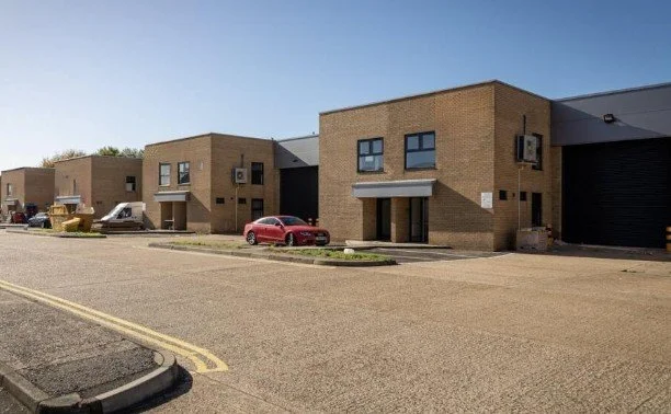 A commercial or industrial building with a brick facade and several parked cars, including a red car, in front of a large parking lot under a clear blue sky.