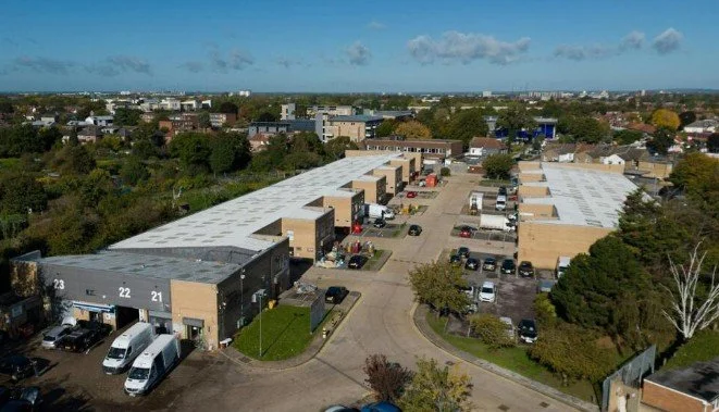 Aerial view of a commercial or industrial building complex with parking lots, surrounded by trees and a city skyline in the background.