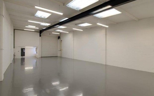 Empty indoor warehouse or industrial space with white walls, gray polished concrete floors, and overhead fluorescent lighting.