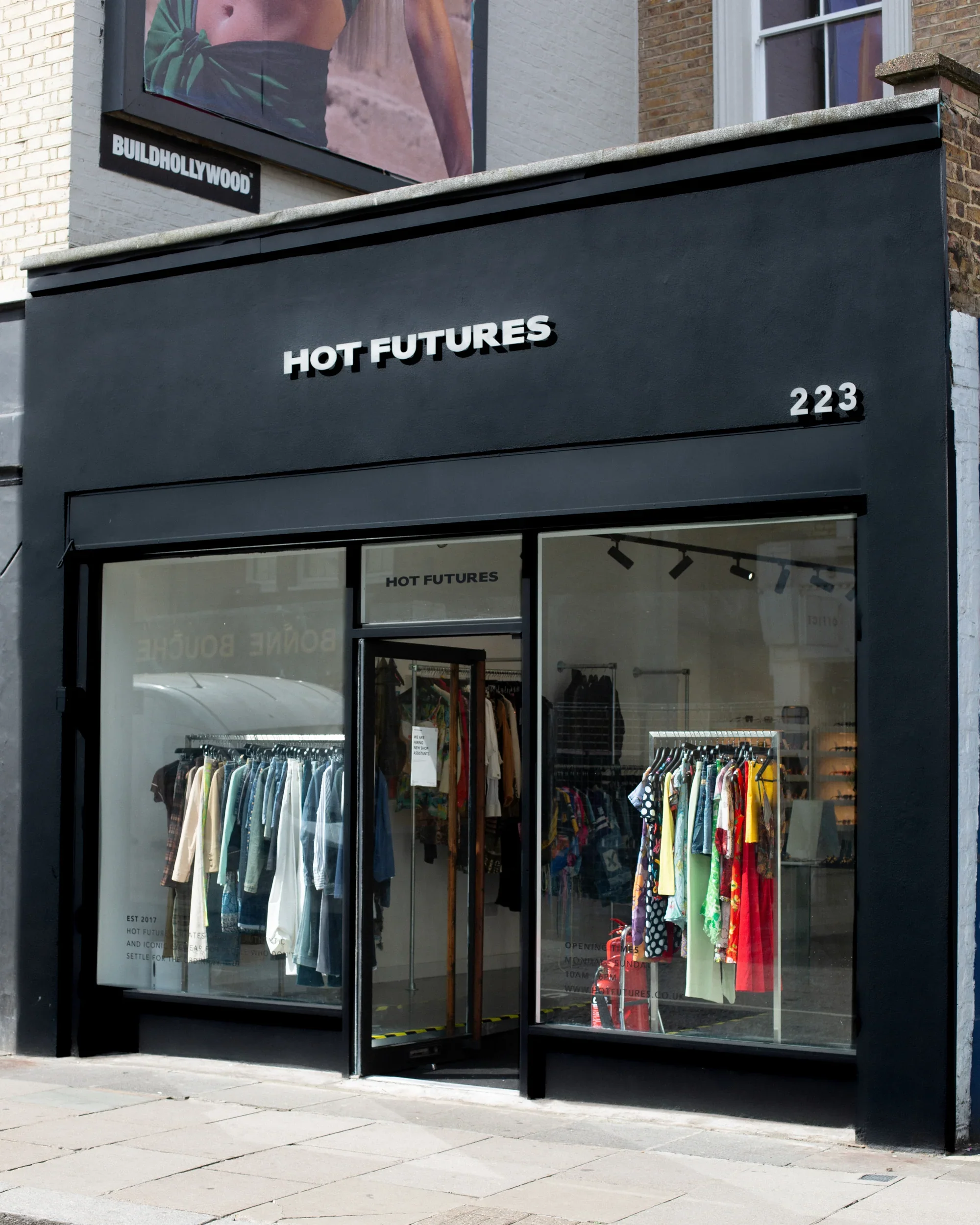 Front view of a clothing store named HOT FUTURES with large glass windows displaying clothing racks, at 223 address, black exterior facade, and a small sign with store name above the door.