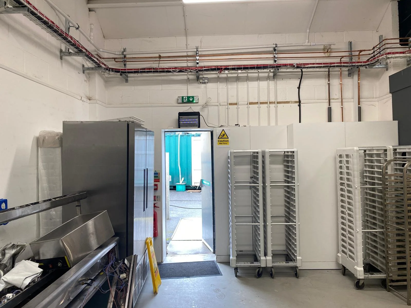 Industrial kitchen or food storage room with metal shelving, carts, and equipment near an open doorway, with visible piping and control panels on the wall.