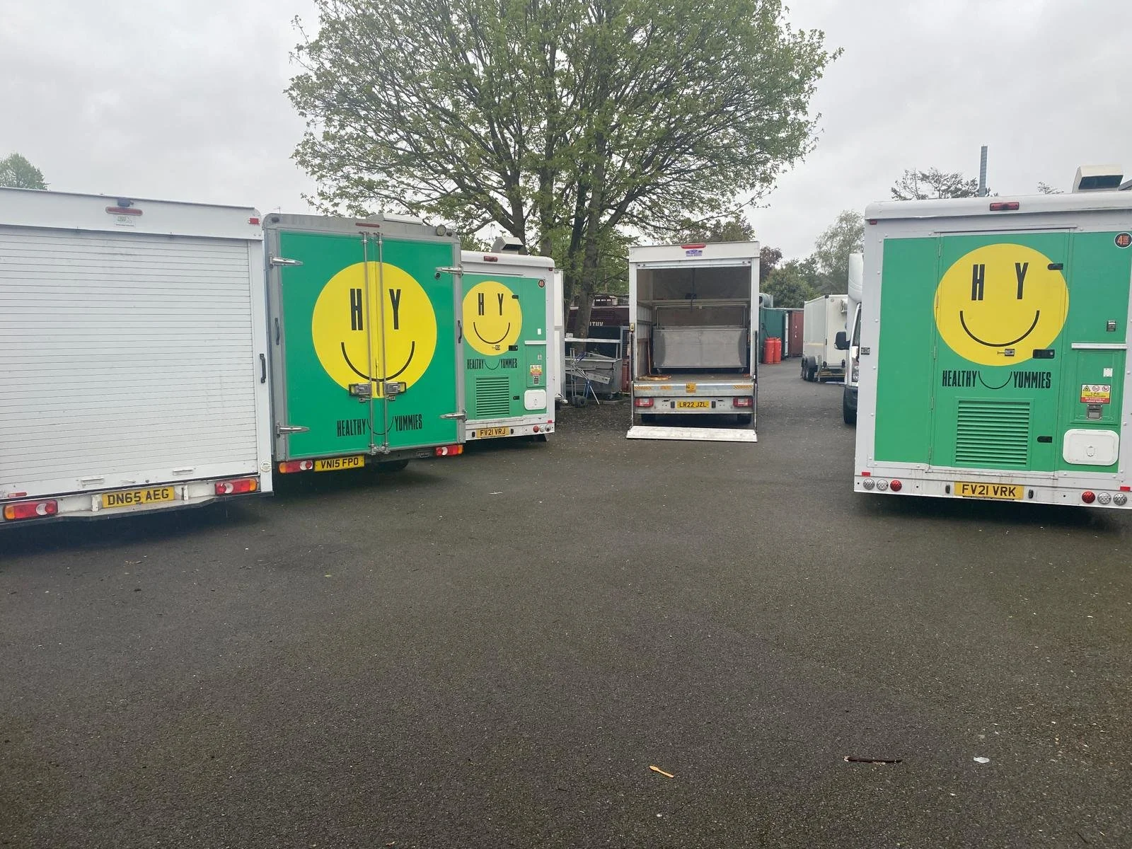 Several food trucks parked on a gray asphalt surface, two of which feature a large yellow smiley face logo with the words 'Healthy Yummies' underneath. A tree with green leaves is visible in the background.