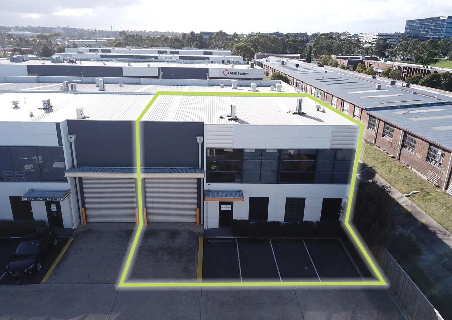 An aerial view of a commercial building with marked boundaries, showing parking spaces, a garage door, and an entrance door, surrounded by other industrial buildings and green areas.