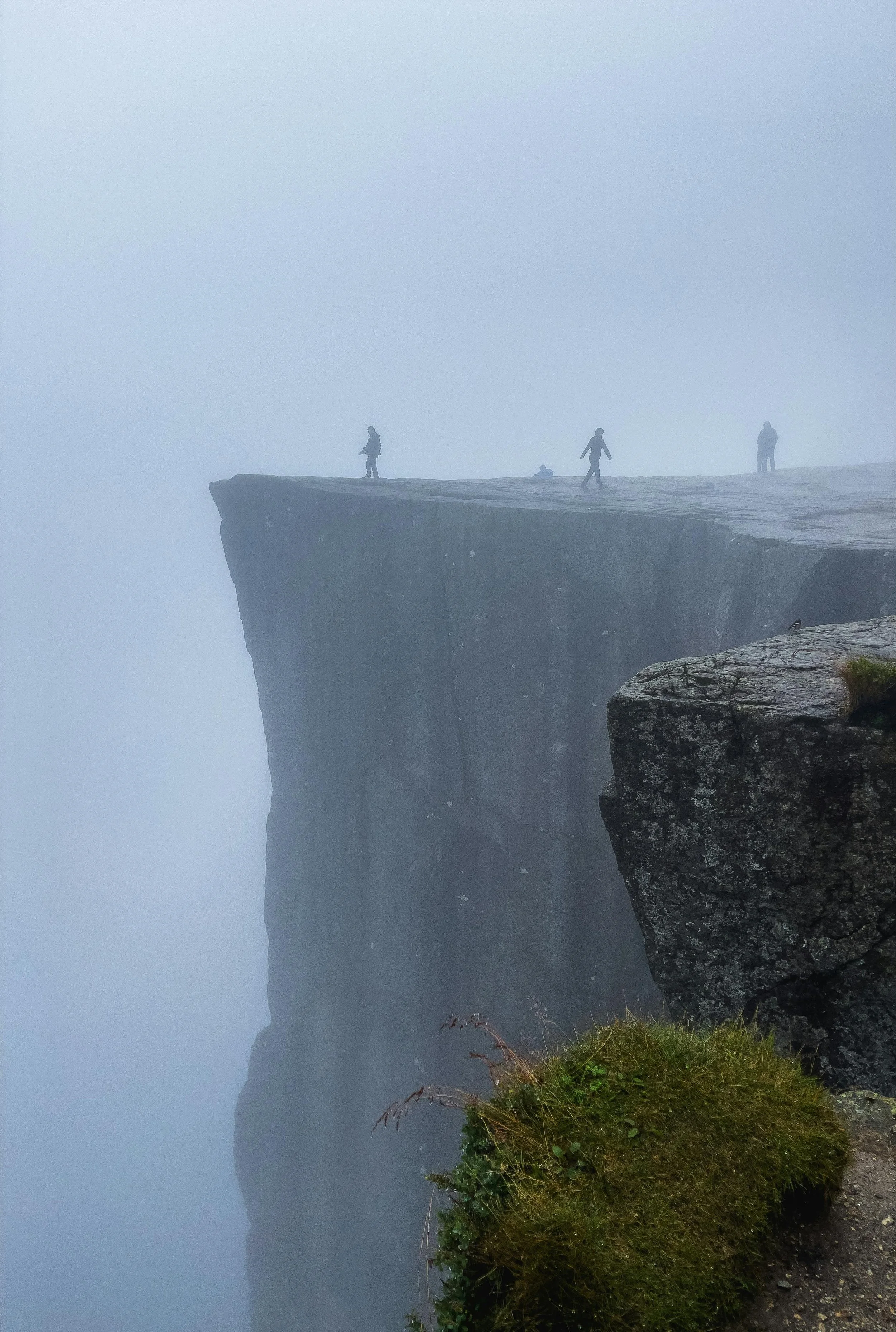 Four people standing and walking on a foggy cliff edge with a sheer drop, with rocks and grass in the foreground.