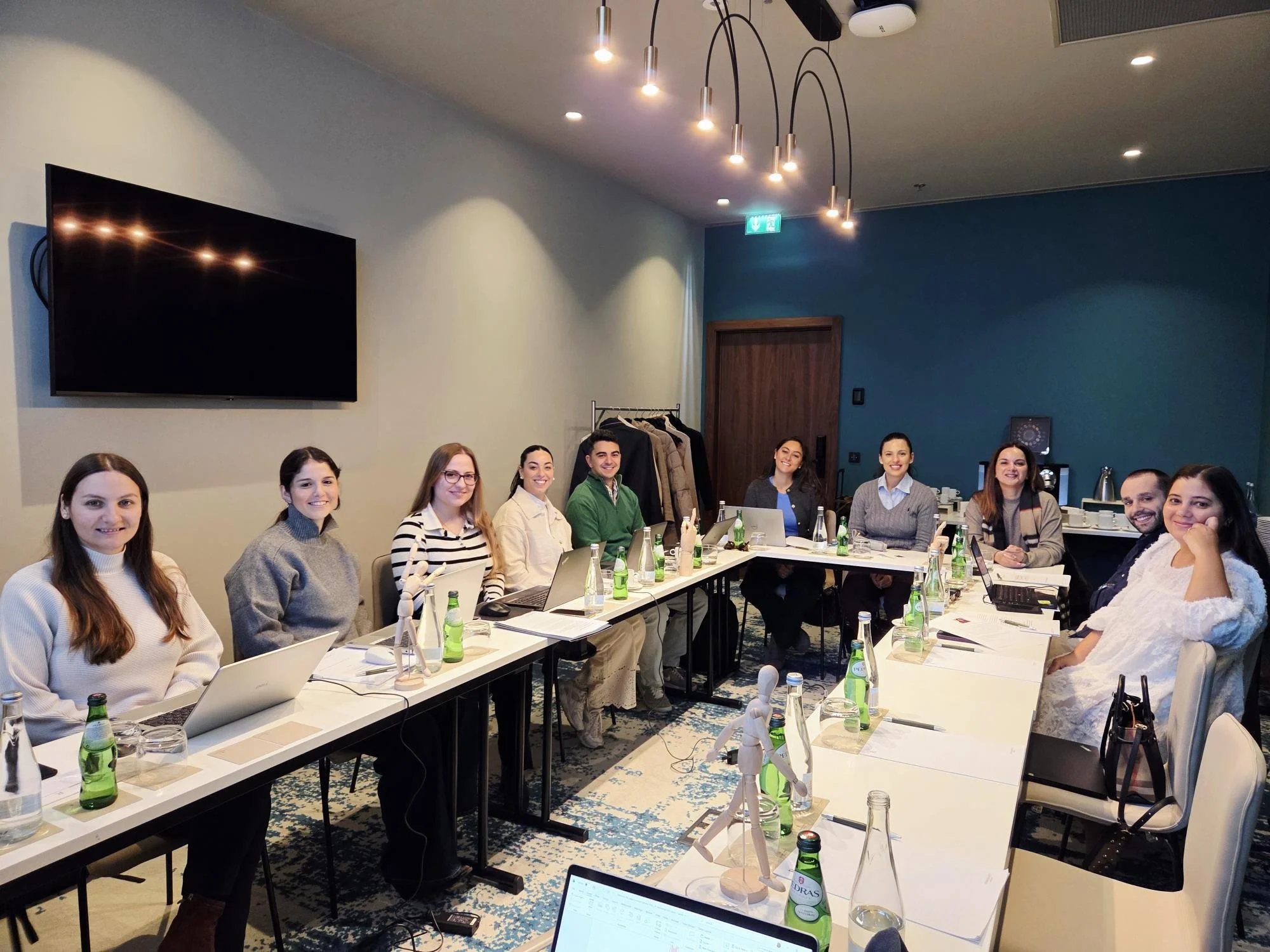 A group of eleven young adults seated around a U-shaped conference table with laptops, notebooks, and bottled beverages in a modern meeting room with a large wall-mounted TV during a legal communications workshop