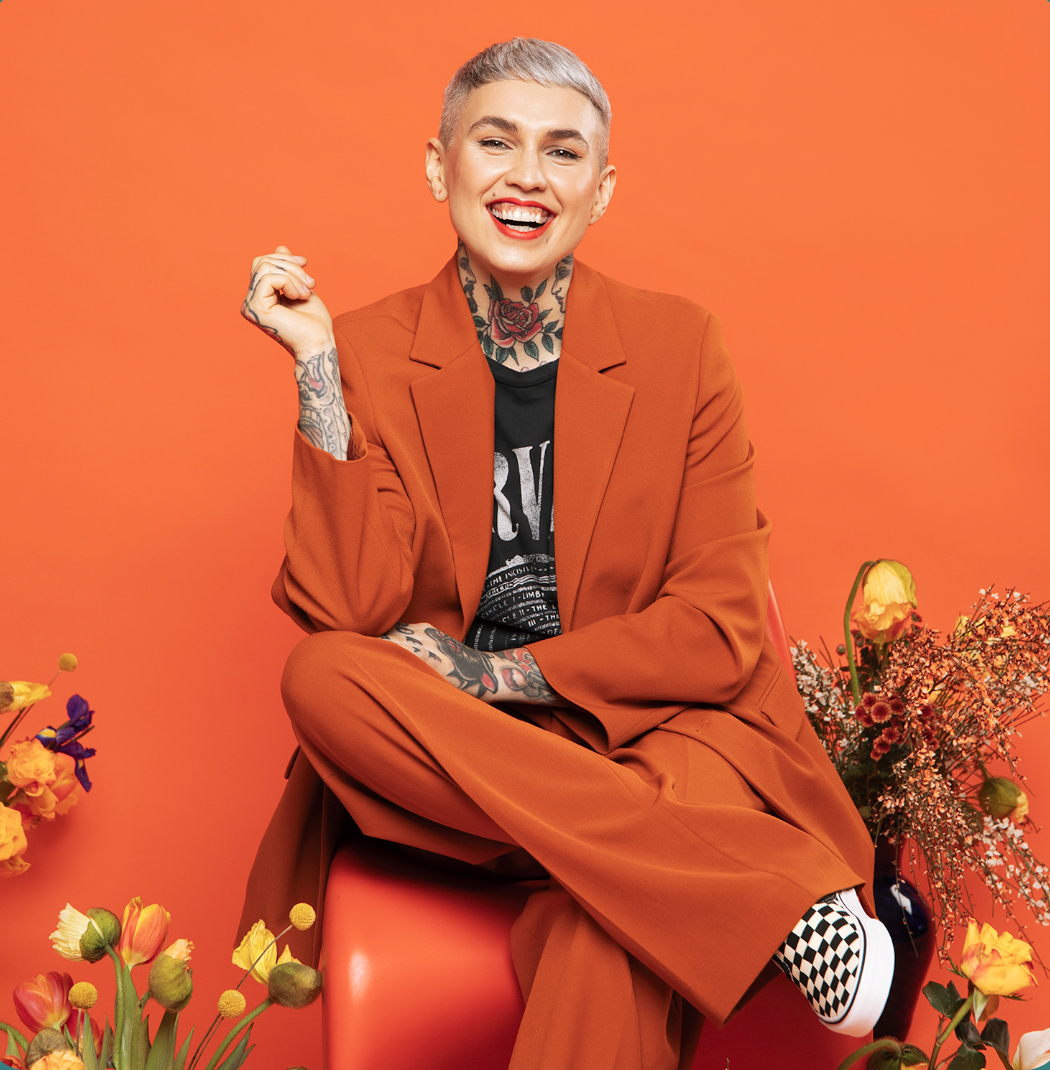A young woman with short gray hair, tattoos on her neck and arms, wearing an orange suit and checkered shoes, sitting on a red chair against an orange background surrounded by colorful flowers, smiling and raising her right hand.