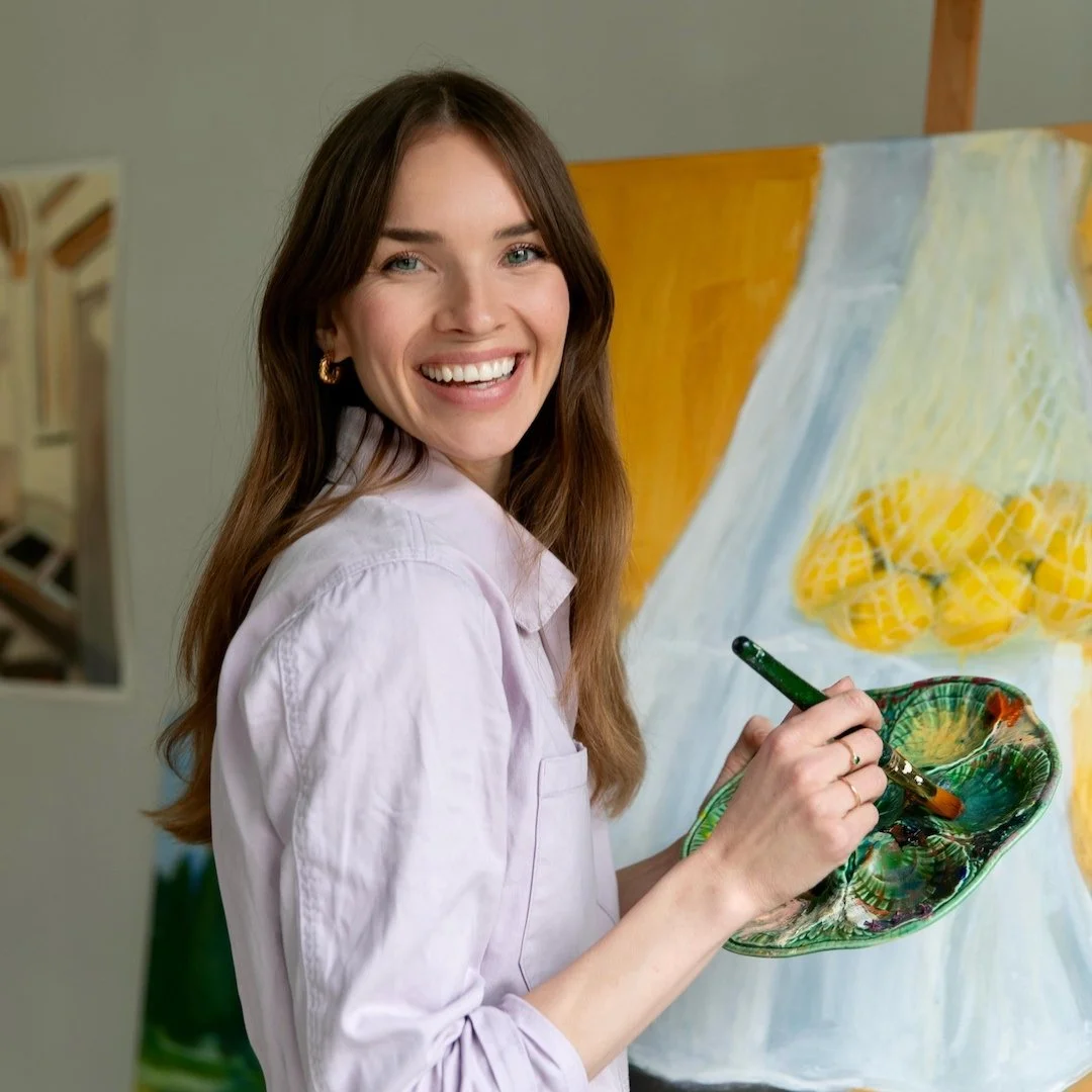A woman smiling while painting on a canvas with a colorful ceramic palette in her hand.