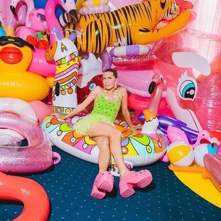 A woman sitting on a colorful inflatable float surrounded by various other inflatables, including animals and cartoon characters, in an indoor setting.