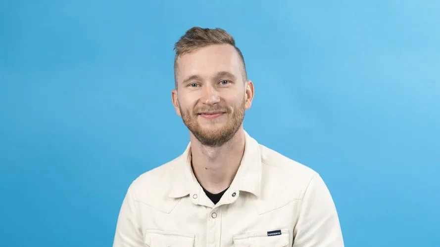 Portrait of a young man with light skin, light brown hair, and a beard, wearing a white jacket, smiling against a blue background.