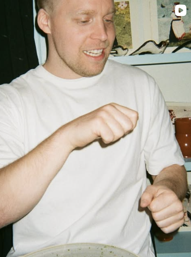 A man in a white t-shirt is looking down and smiling with his fists slightly clenched, appearing to be excited or happy.