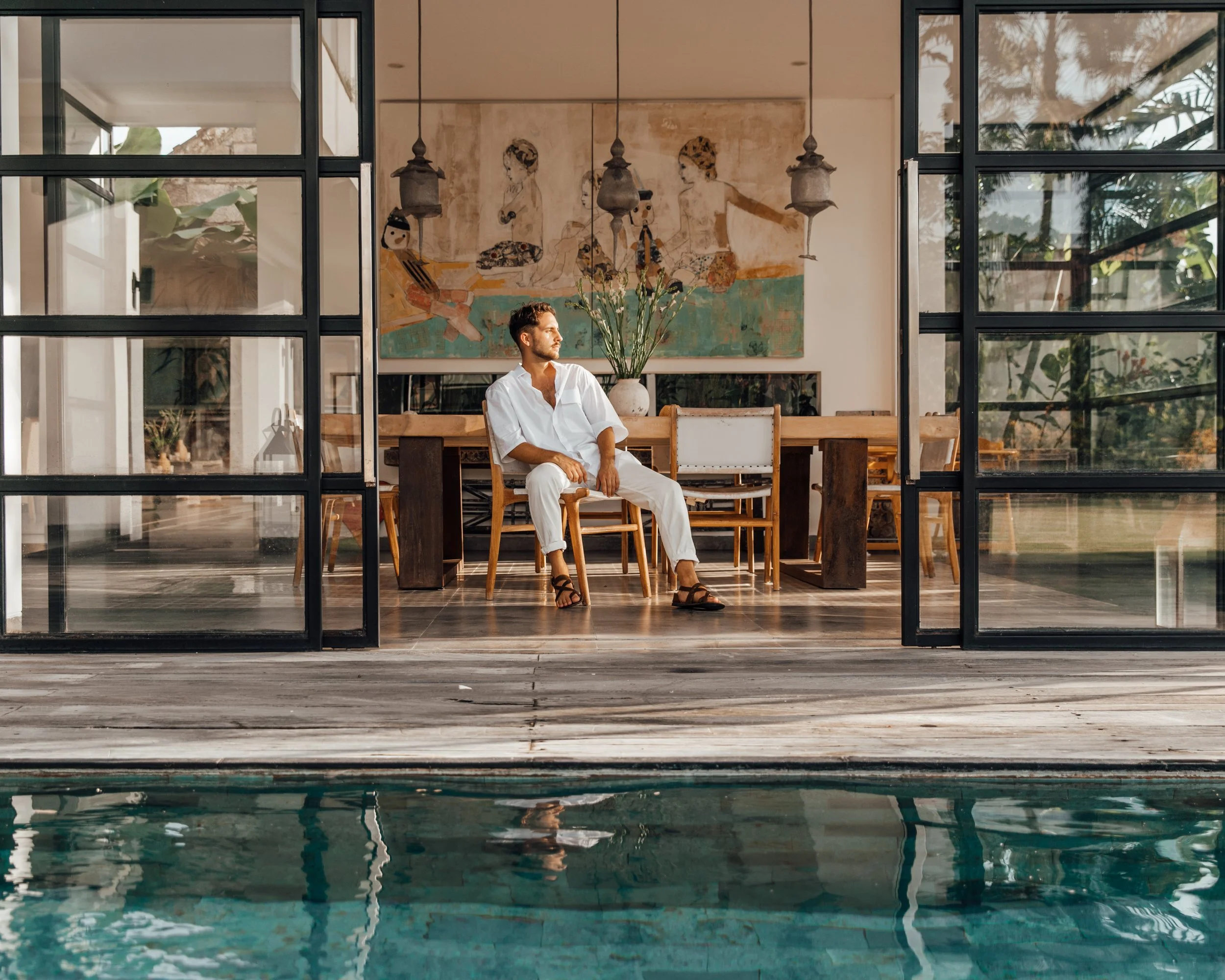 A man in white clothes sitting on a chair inside a house, looking outside through glass doors, with a swimming pool visible in the foreground.