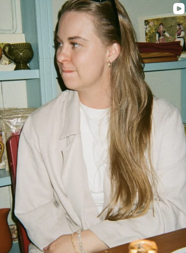 A woman with long, wavy blonde hair sitting indoors, wearing a white jacket and earrings, looking to her left.