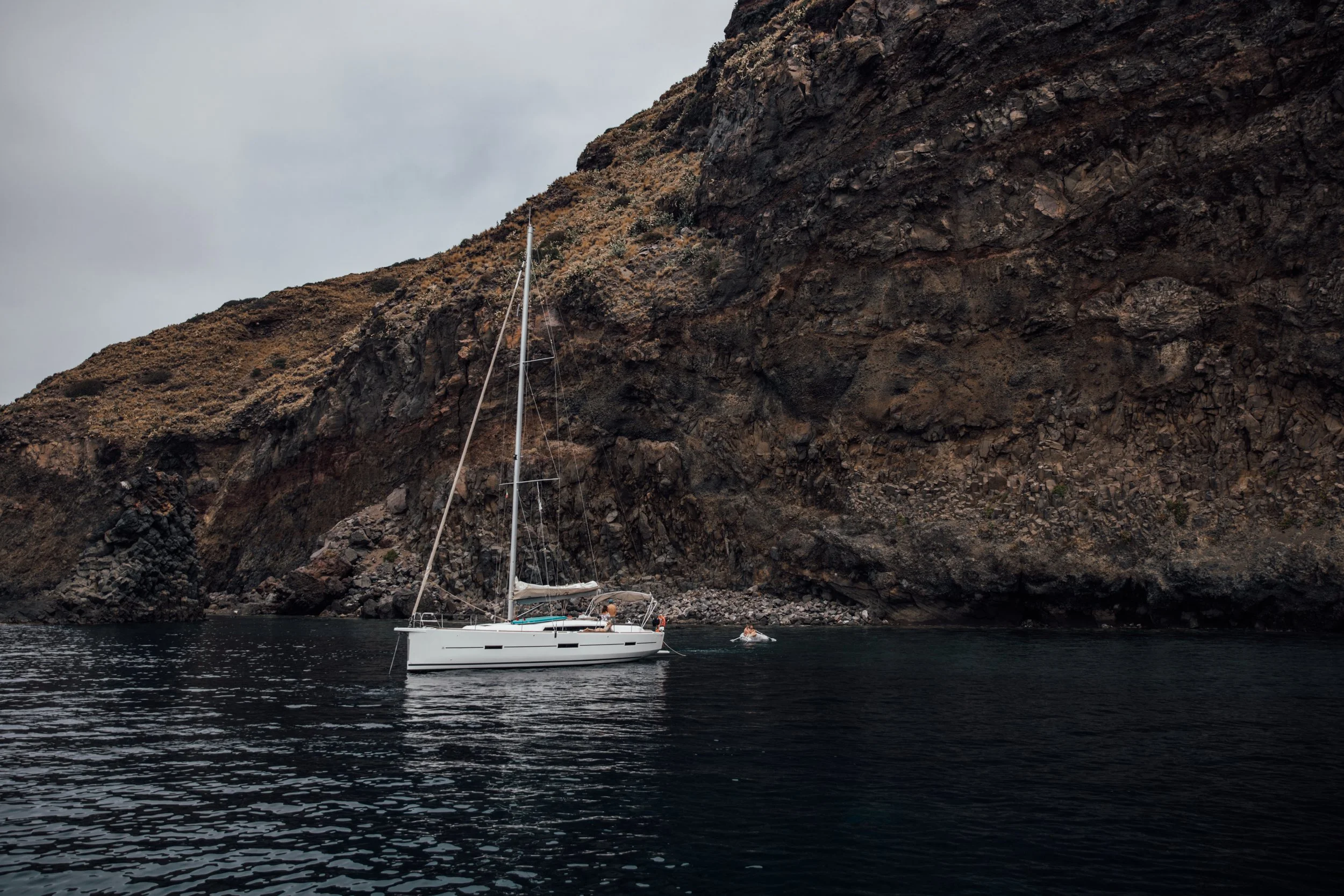 A sailboat anchored near a rocky cliffside with a person on the deck and a small motorboat nearby.