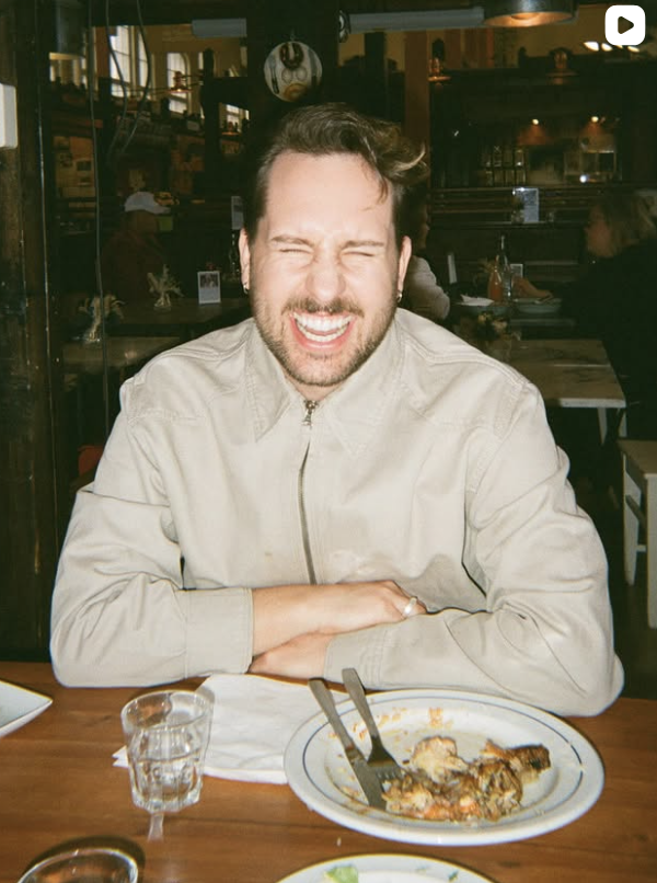 A man with short dark hair and a beard is sitting at a restaurant table, smiling with his eyes closed. He is wearing a light-colored zip-up jacket. There is a partially eaten plate of food and a glass of water in front of him.