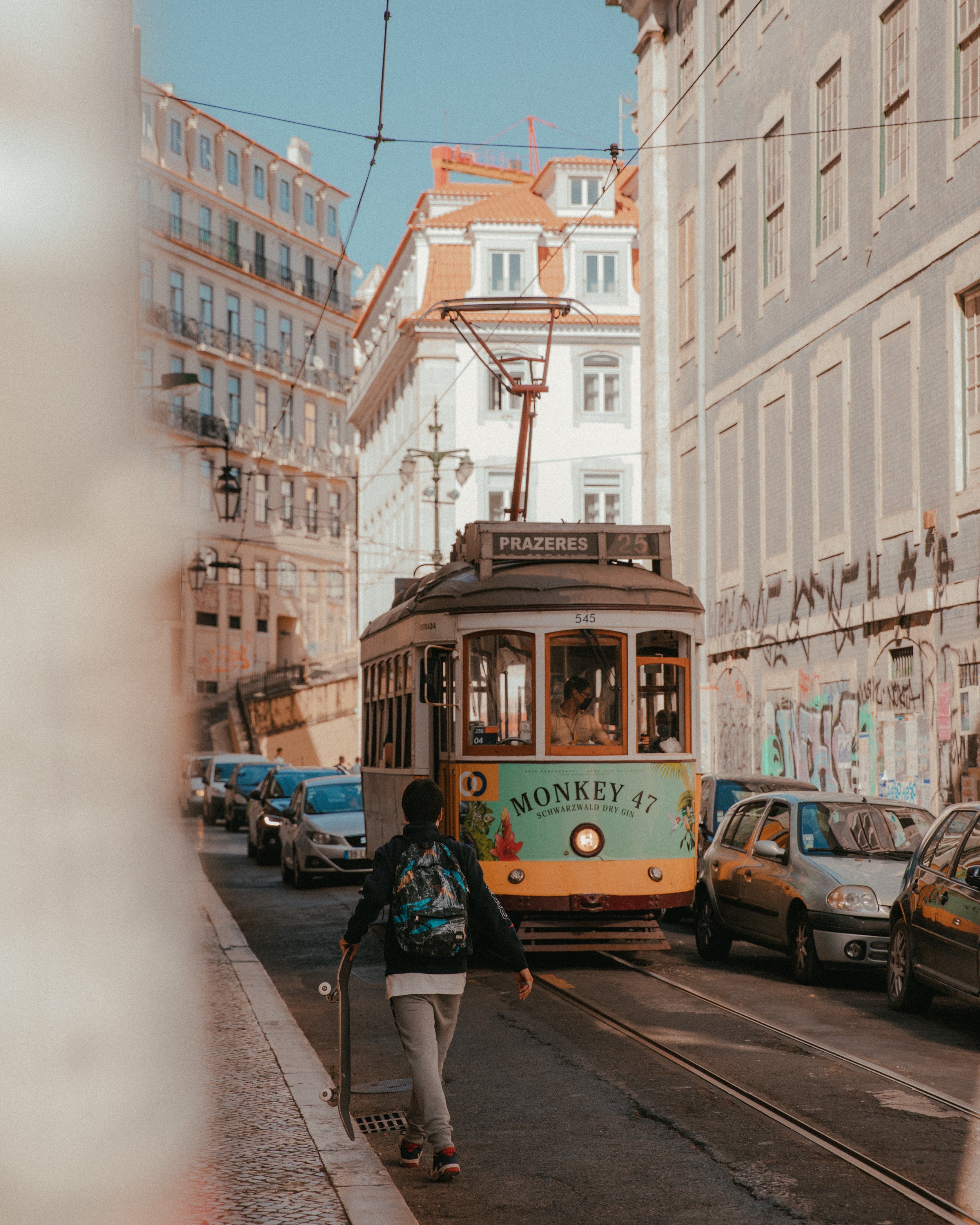 A vintage tram approaching on a city street with cars parked along the side, graffiti on the walls, and a pedestrian walking with a skateboard.