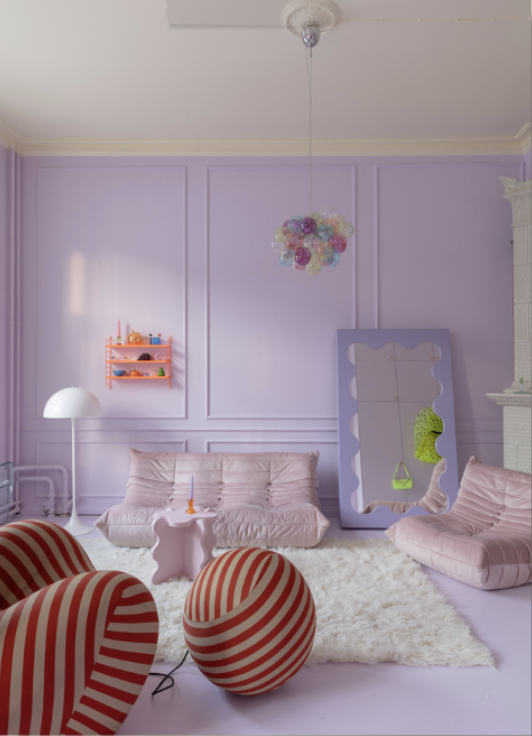 A pastel purple living room with various furniture including a pink couch, a pink armchair, and a pink side table. There is a white shaggy rug on the floor, a large mirror with a purple frame leaning against the wall, and a colorful chandelier hanging from the ceiling. A small pink shelf with toys and a white floor lamp are also visible.