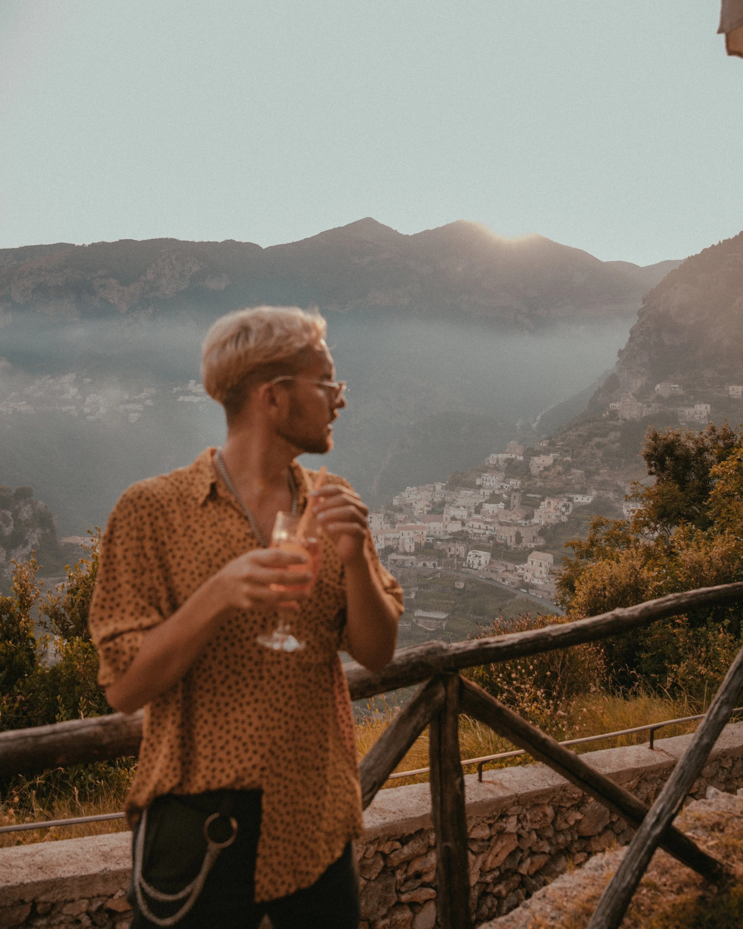 A man with light hair and glasses holding a drink, standing on a scenic overlook with mountains and a hillside village in the background at sunset.