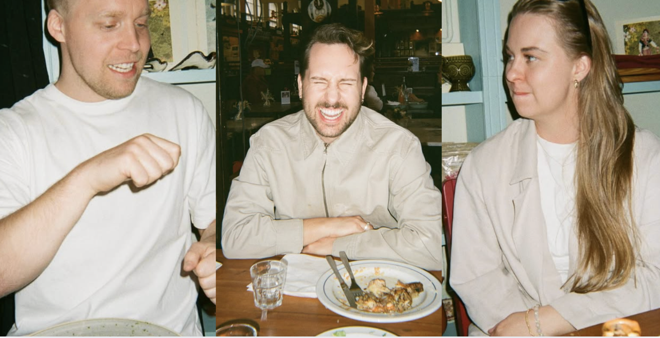 Three people at a restaurant table. The man in the middle is smiling with his eyes closed, with a partly eaten plate of food in front of him. The man on the left is looking down with a raised fist. The woman on the right is looking at the man in the middle with a slight smile.