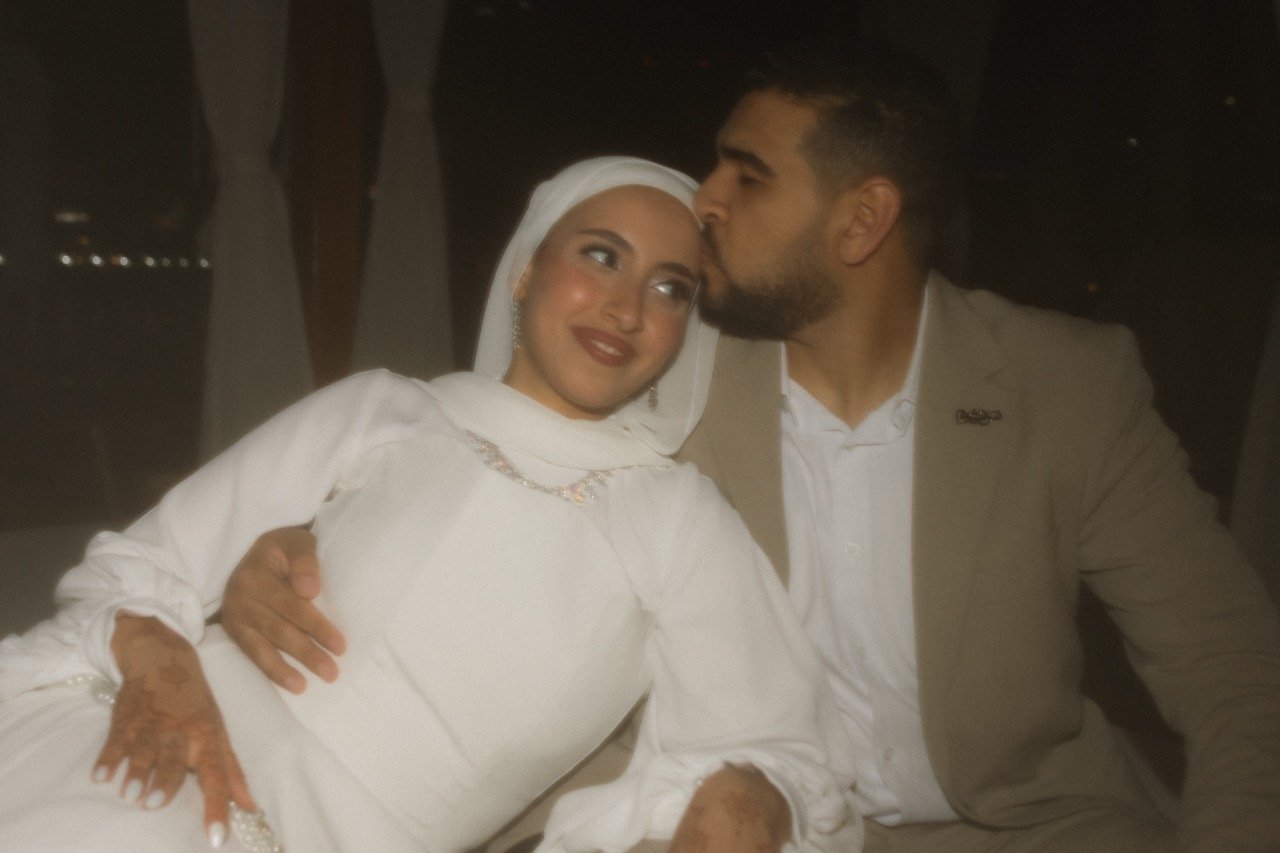A woman wearing a white dress and headscarf receives a kiss on the forehead from a man in a beige suit at a dimly lit event, possibly a wedding.