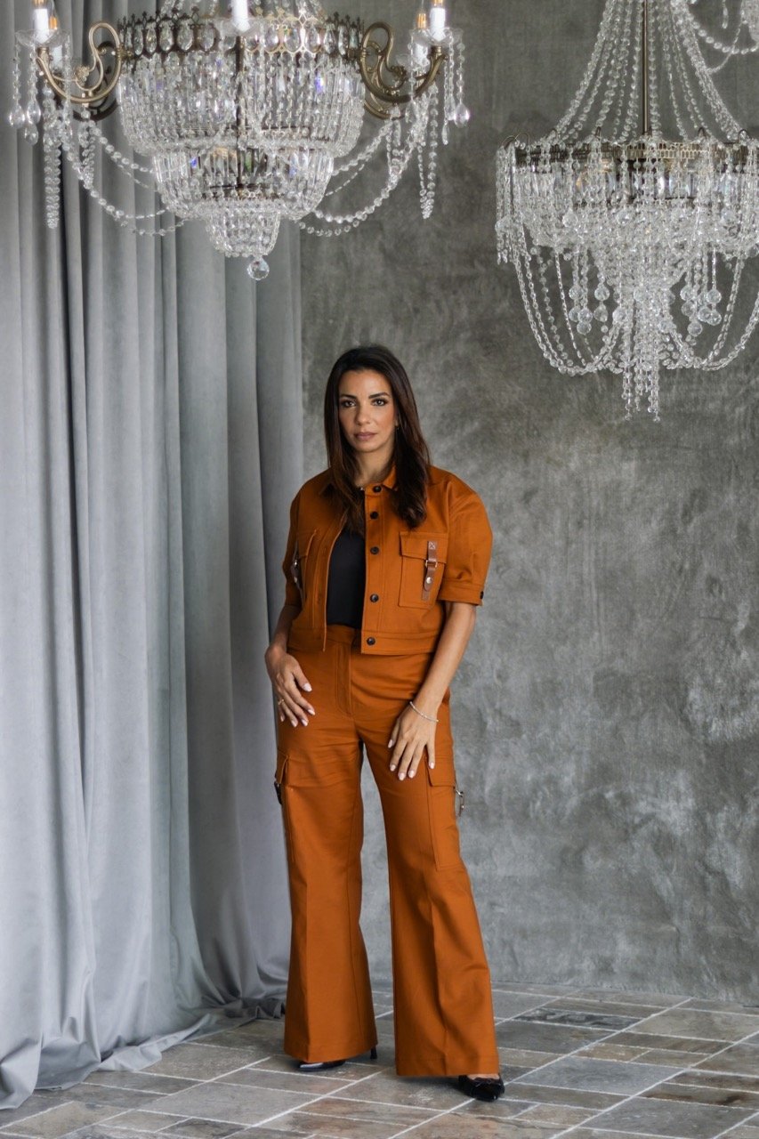 A woman stands in front of a gray wall with two large crystal chandeliers hanging above her. She is wearing a rust-colored coordinated outfit with a cropped jacket and wide-legged pants, and has long dark hair.