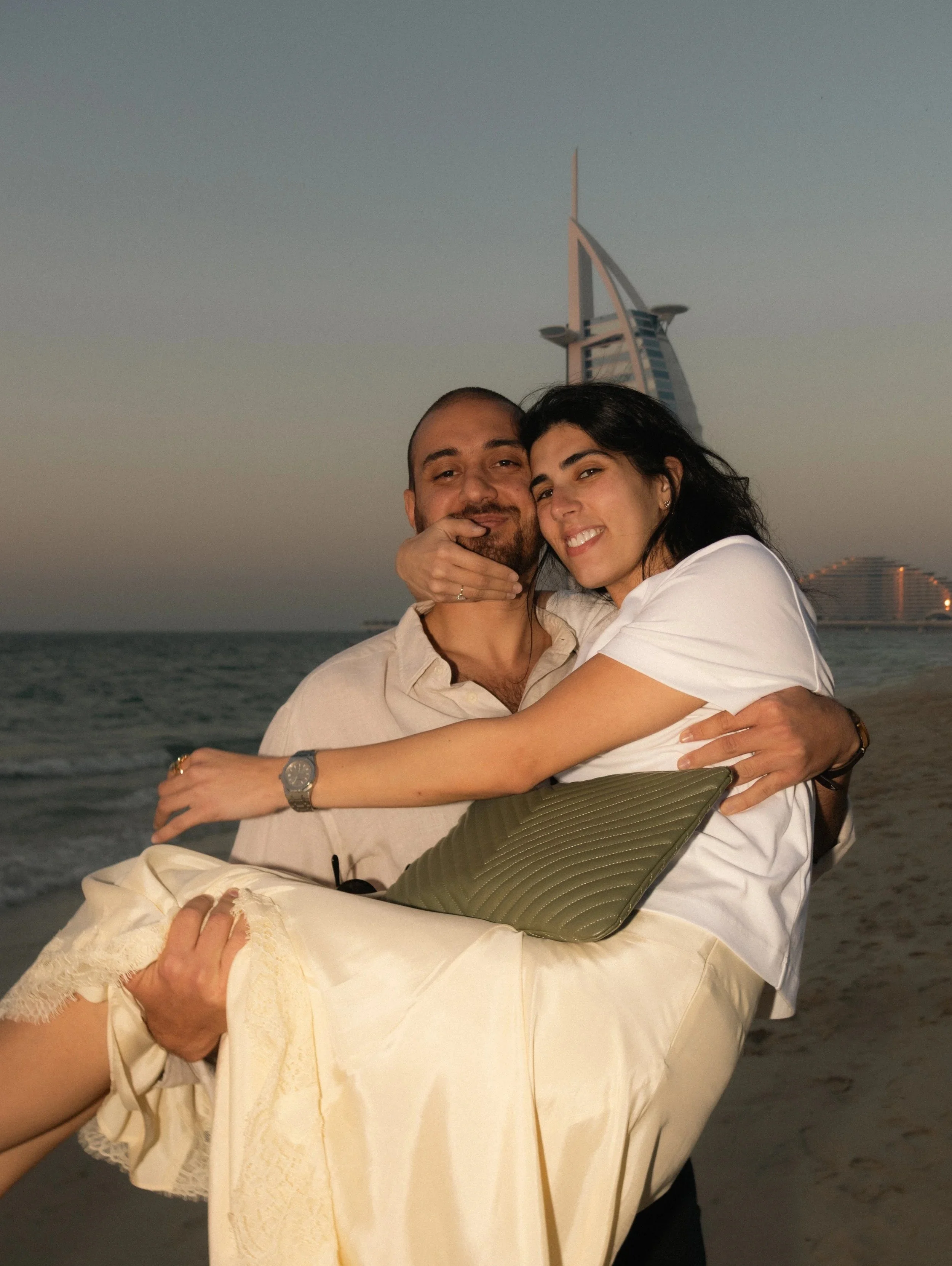 A couple hugging each other on a beach with the Burj Al Arab hotel in Dubai in the background, during sunset.