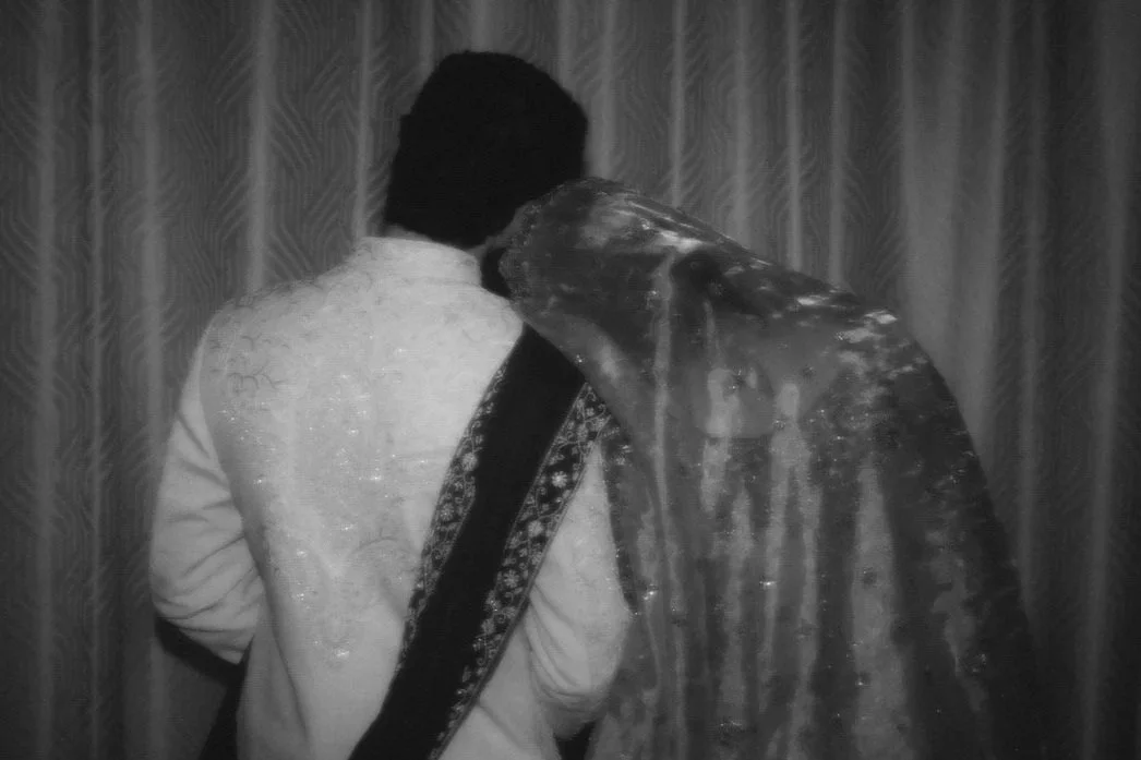 Man and woman in traditional wedding attire sharing a cake at their wedding reception, with beige draped backdrop and floral arrangements.