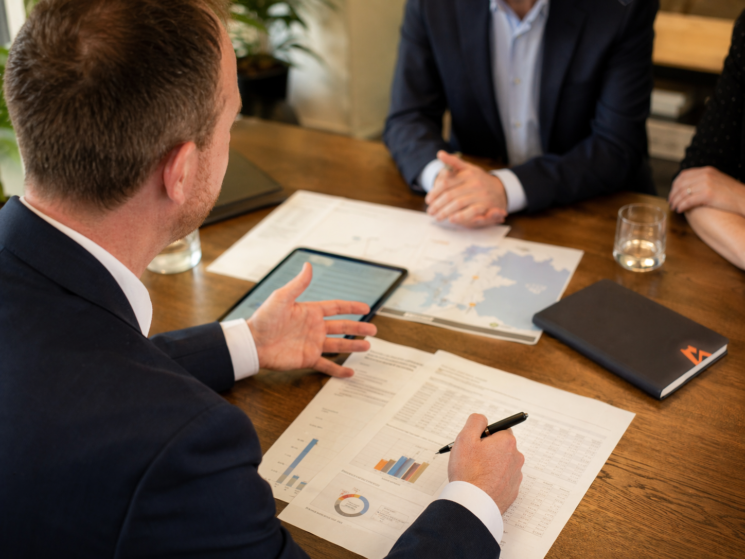 Business meeting with three people, one man speaking and others listening, documents and graphs on the wooden table, a tablet, a notebook, and a glass of water visible.
