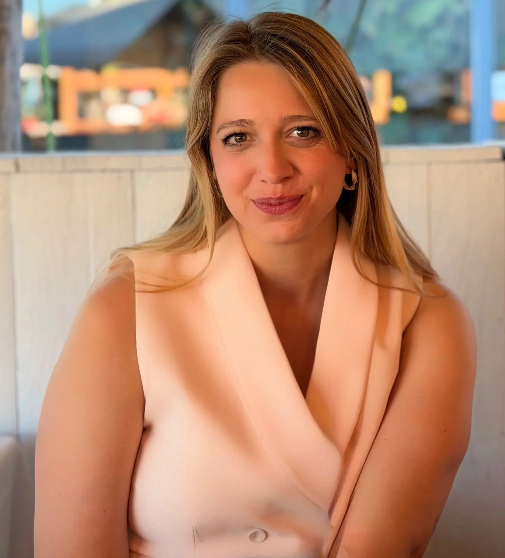 Nahaufnahme einer lachenden Frau mit blondem Haar, die sitzt, im Hintergrund ist ein Zaun und Häuser sichtbar.