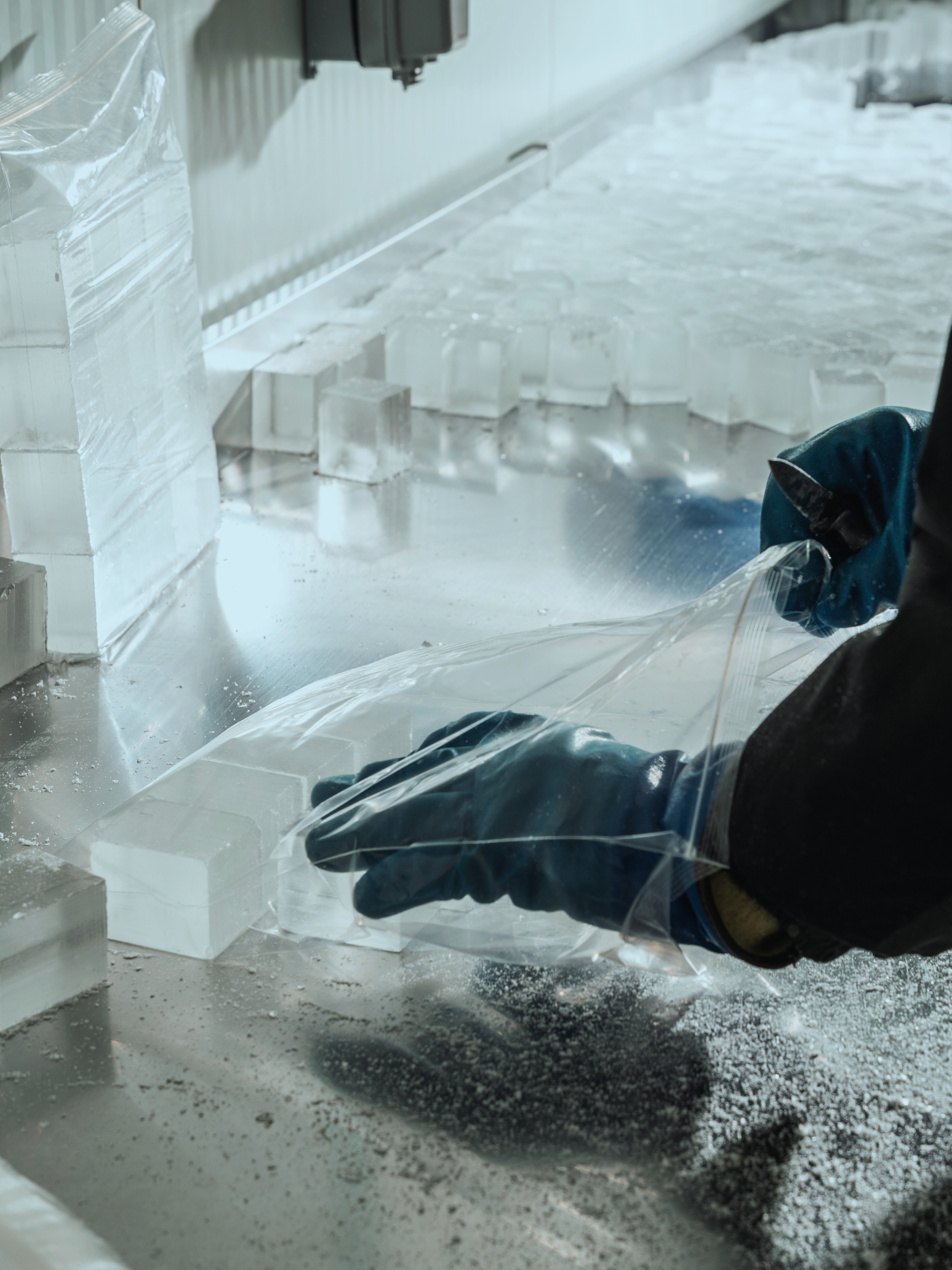 Person wearing black gloves and a dark jacket handling a plastic bag of ice on a stainless steel surface in a commercial kitchen or ice manufacturing facility.