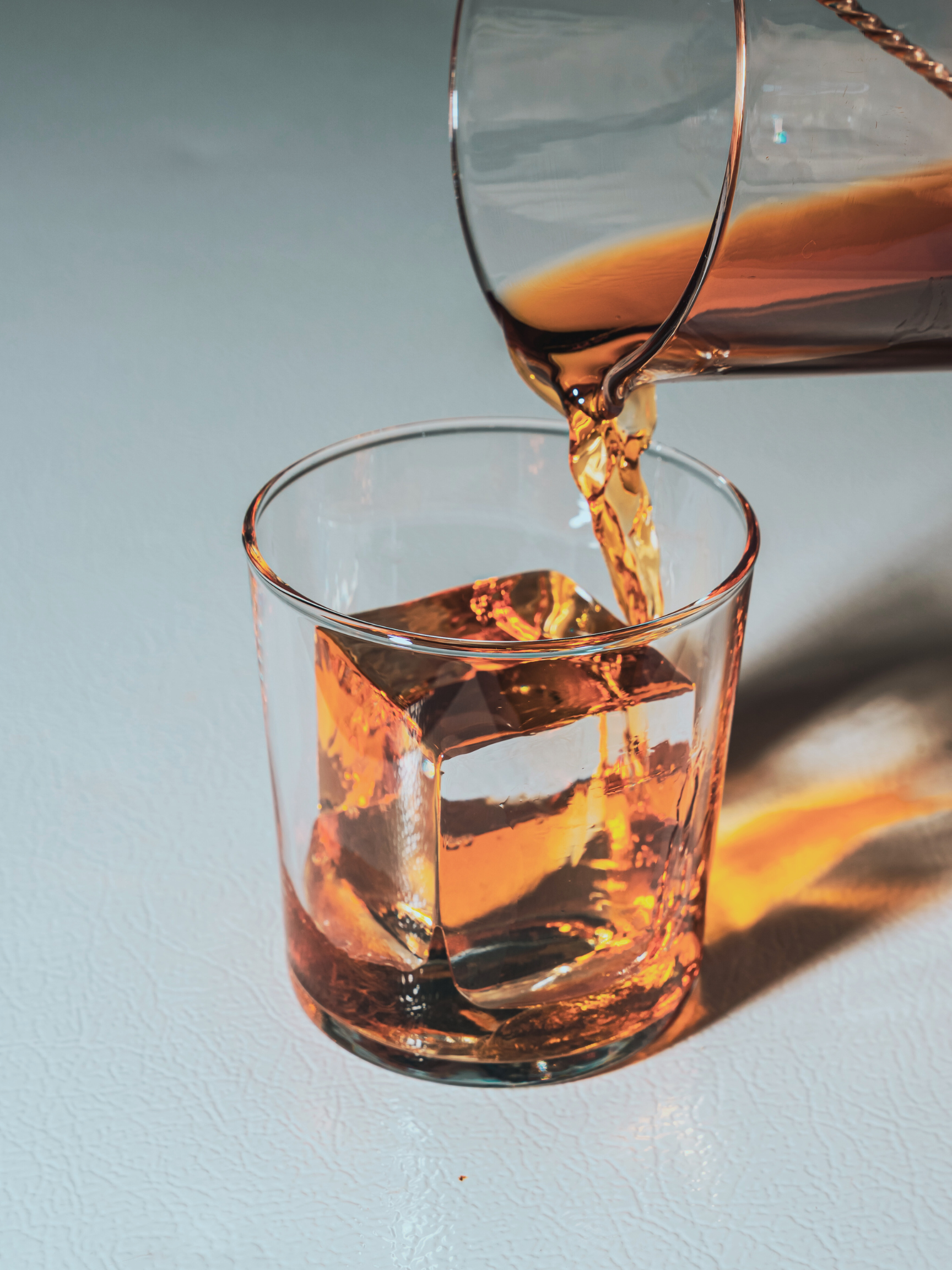 A glass with clear ice cube and a copper-colored liquid being poured into it from a pitcher.