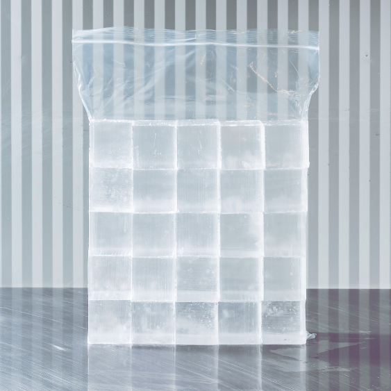 A large clear plastic bag filled with ice cubes, placed on a stainless table with a striped wall in the background.