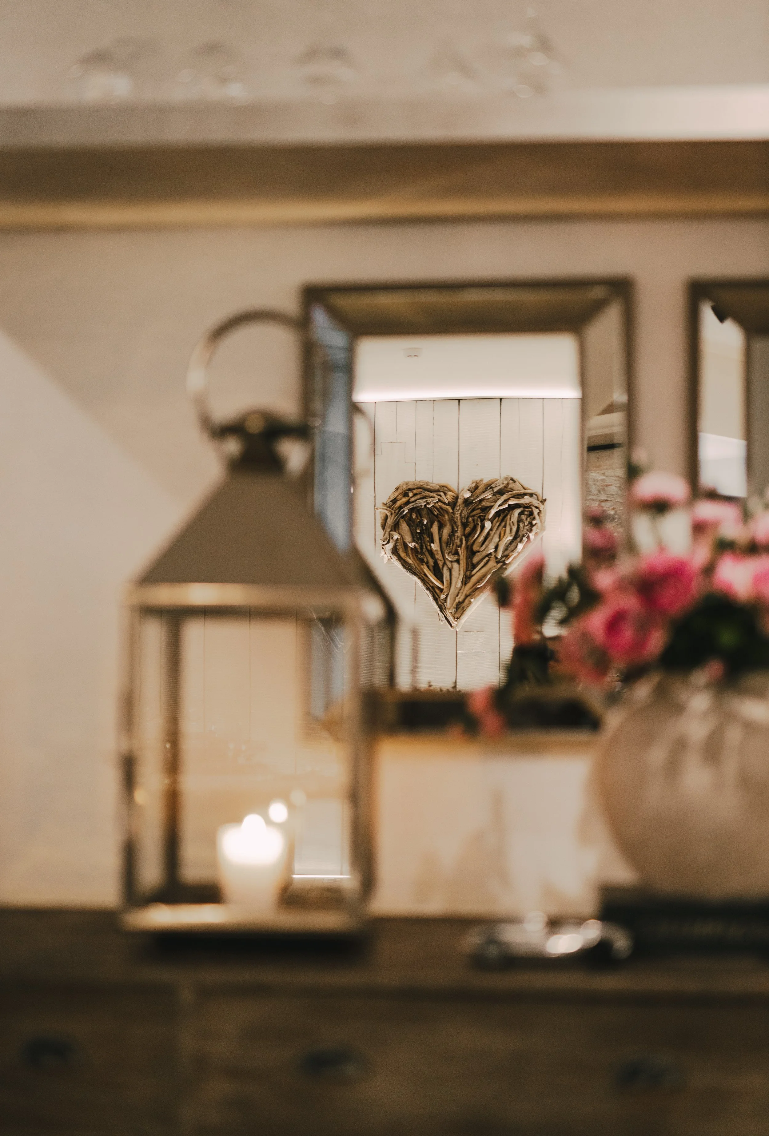 A wooden heart-shaped decoration hangs on a white wooden wall, reflected in a mirror. In the foreground, there is a lantern with a lit candle and a bouquet of pink flowers.
