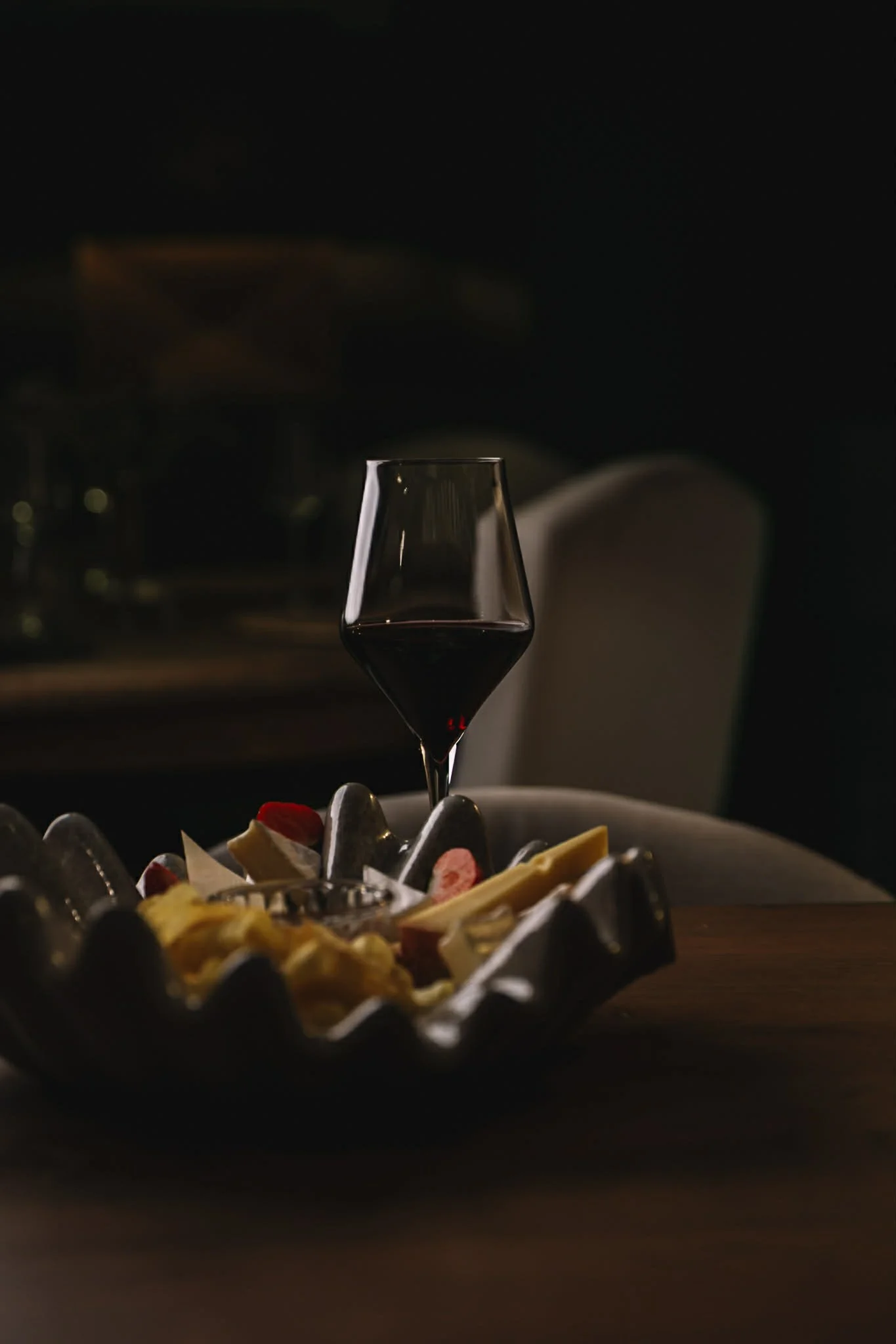 A glass of red wine on a wooden table. A decorative bowl containing chocolates and strawberries is in front of the glass. The background is dark and out of focus.