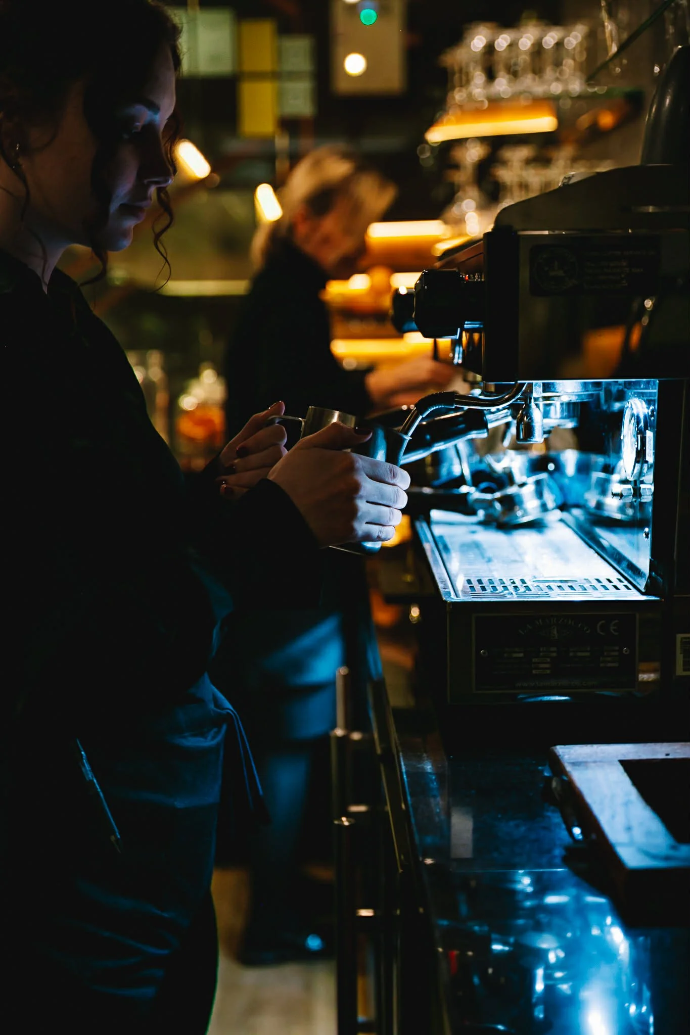 A barista preparing a coffee with an espresso machine in a dimly lit cafe, with another employee working in the background.