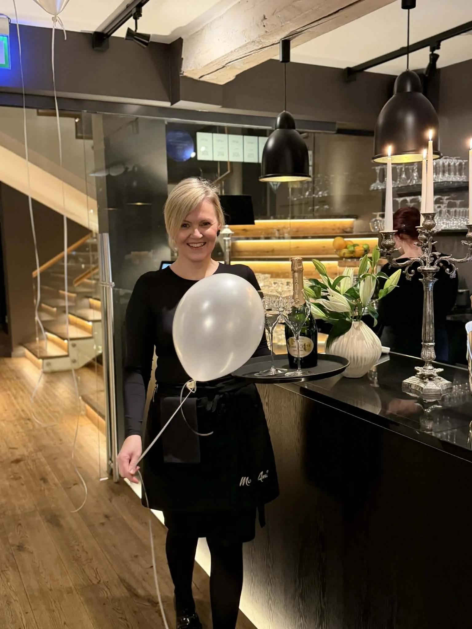 A woman in a black dress and apron toasting with a tray of champagne glasses and bottle in a modern restaurant or bar setting with candles, flowers, and black pendant lights.