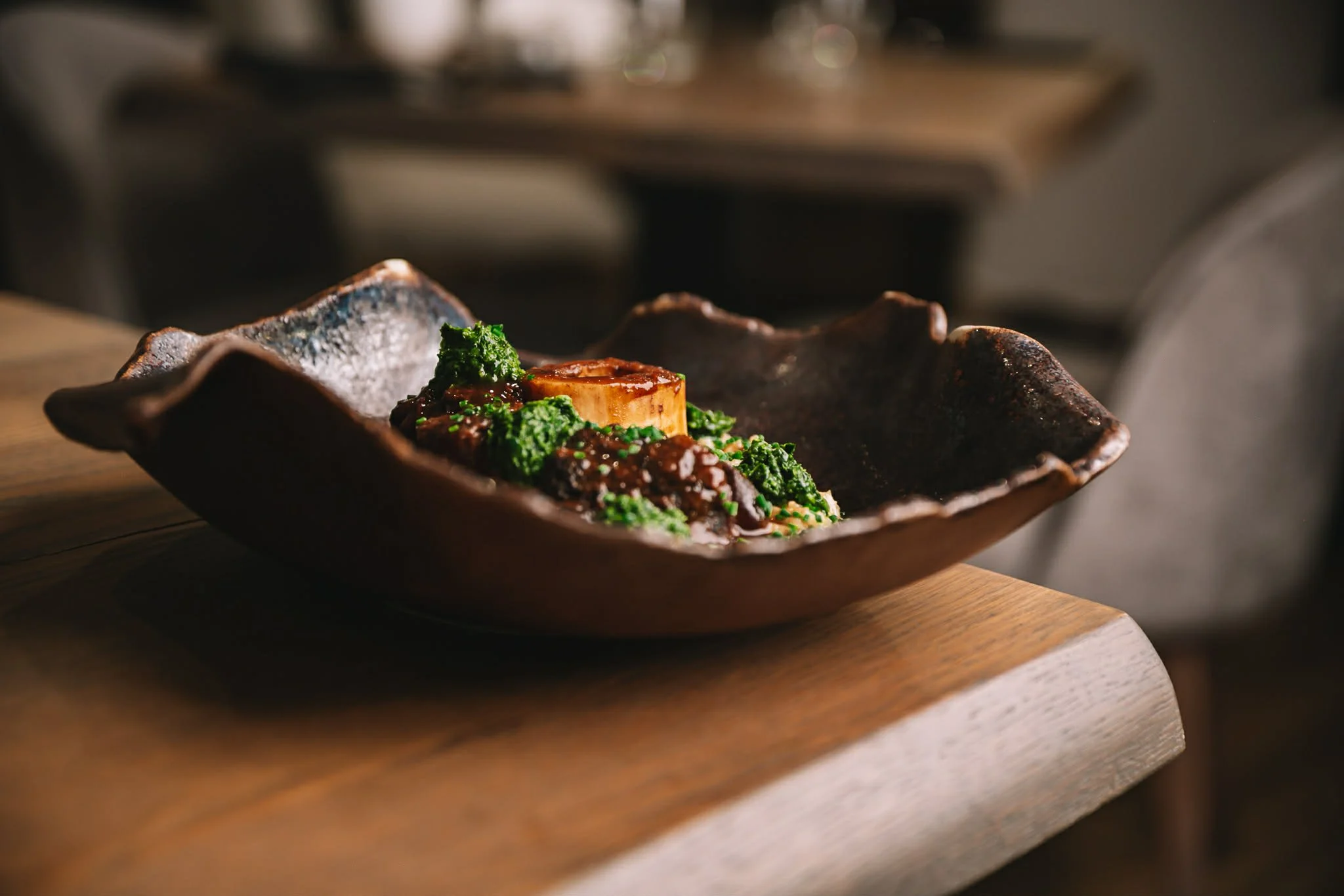 A dish of cooked meat topped with green vegetables and a round slice of root vegetable, served in a rustic, wavy-edged ceramic bowl on a wooden table.