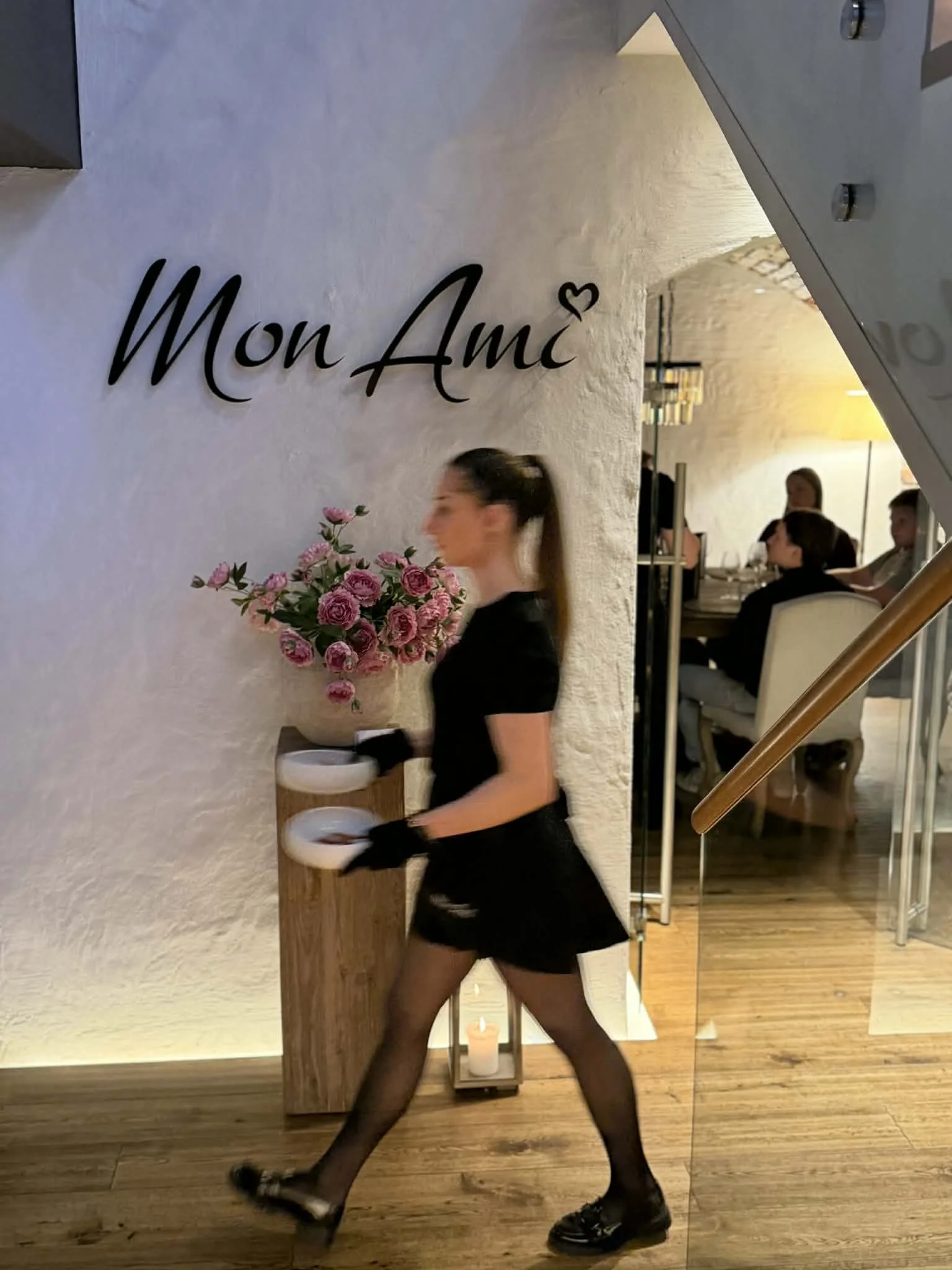 A woman dressed as a server walking past a wall with 'Mon Amie' written on it and a heart symbol, holding a tray inside a restaurant.
