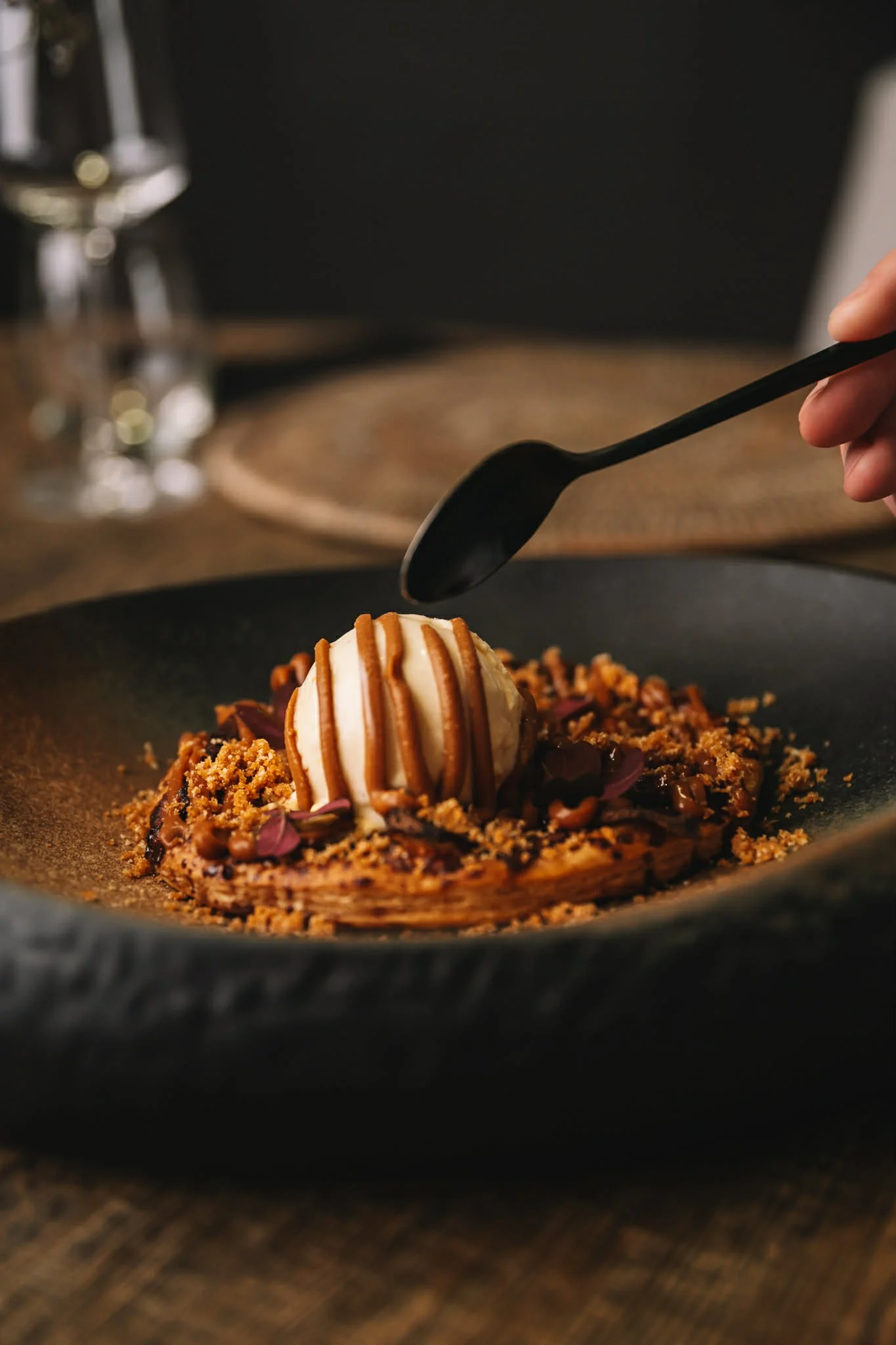 A dessert with a scoop of vanilla ice cream drizzled with caramel sauce on a bed of crumbled cookie or biscuit, topped with pink edible flowers, served on a dark plate.