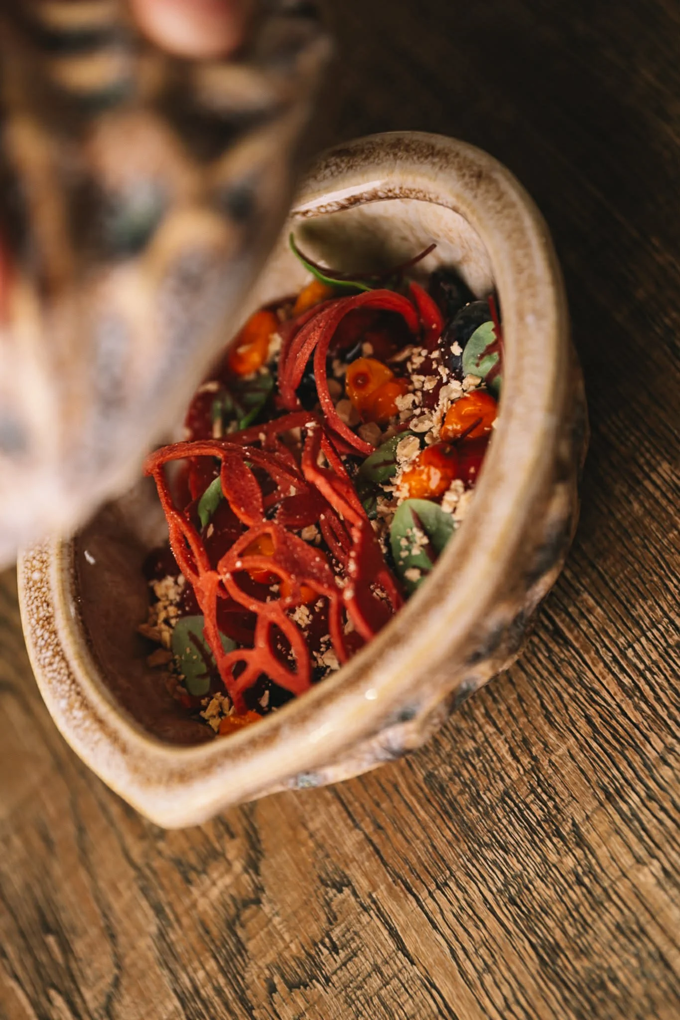 A rustic bowl containing a colorful salad with shredded red vegetables, cherry tomatoes, green herbs, and crushed nuts or seeds on a wooden surface.