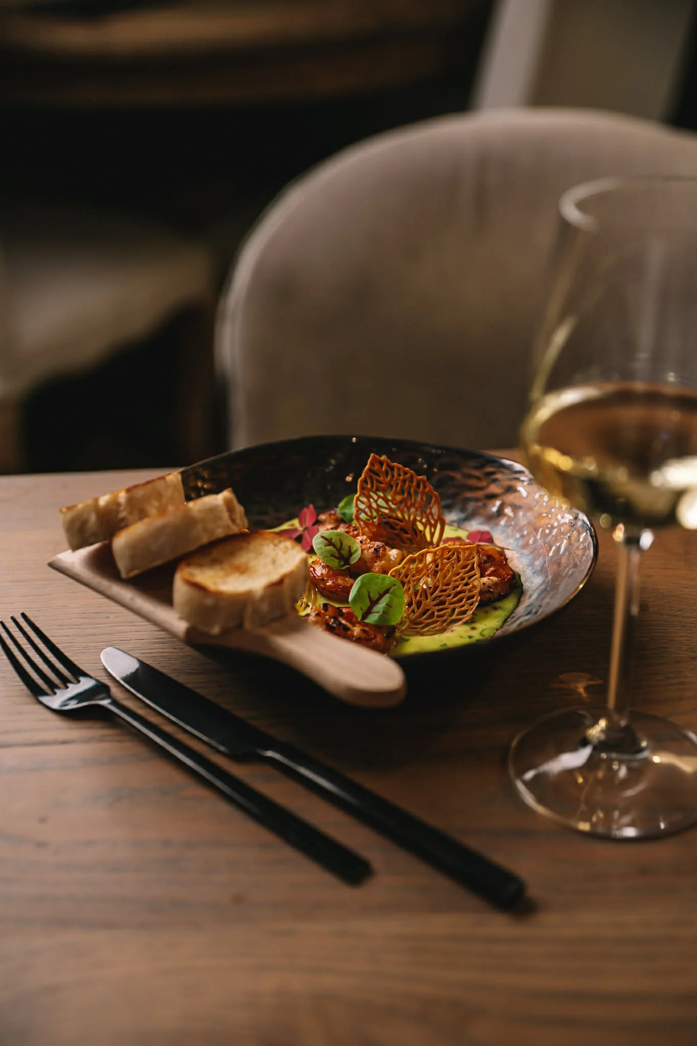 A plate of gourmet food with toasted bread, grilled vegetables, and decorative carrot chips, garnished with microgreens, on a wooden table with a glass of white wine.