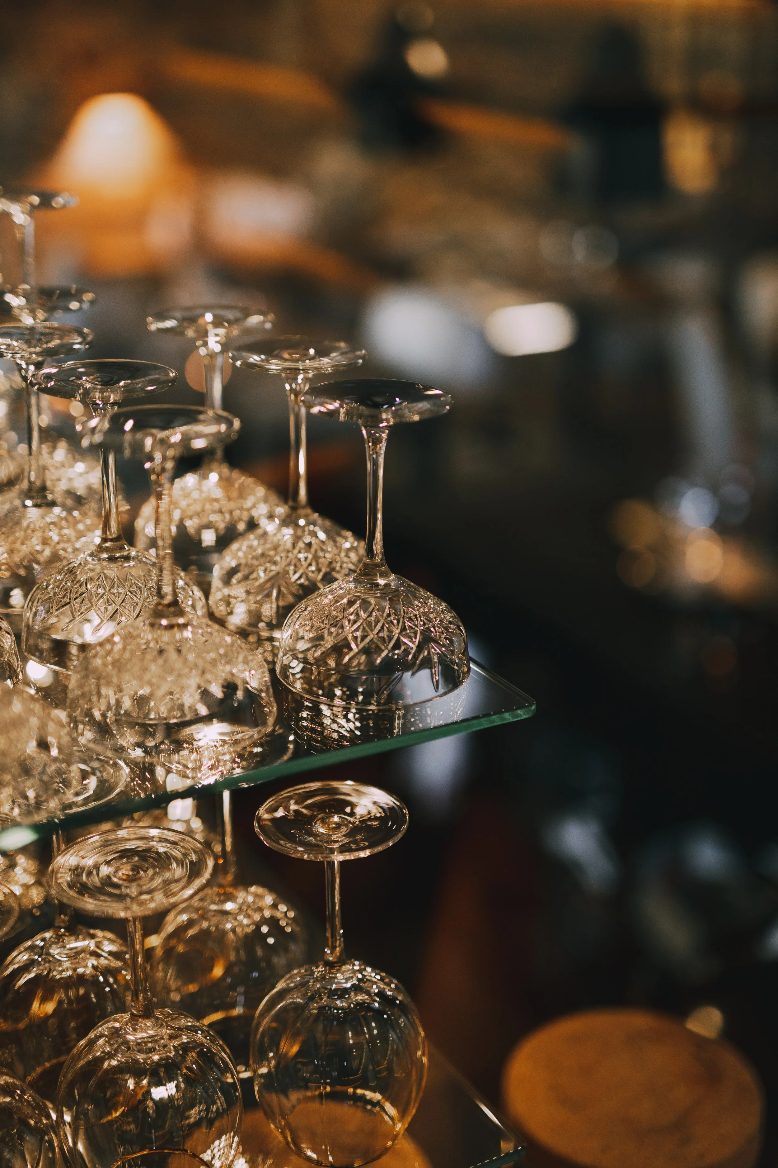 A collection of upside-down crystal wine glasses on a glass shelf with a warm, blurred background.